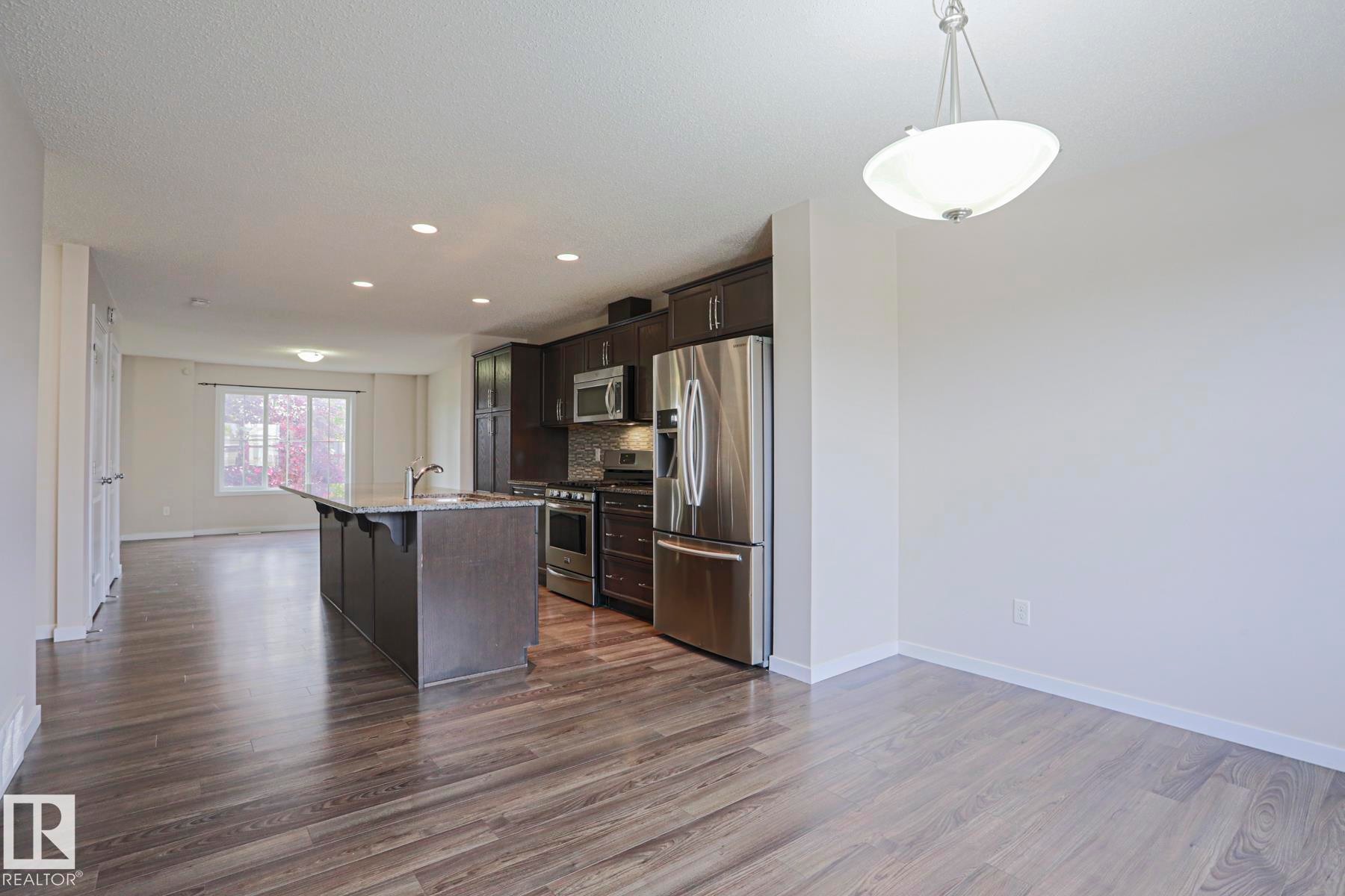 Open-concept layout with wood-finish flooring, dark wood cabinetry, stainless steel appliances, and a central kitchen island with seating - 308 Watt Boulevard Sw, Edmonton, AB - Indoor Photo Showing Kitchen