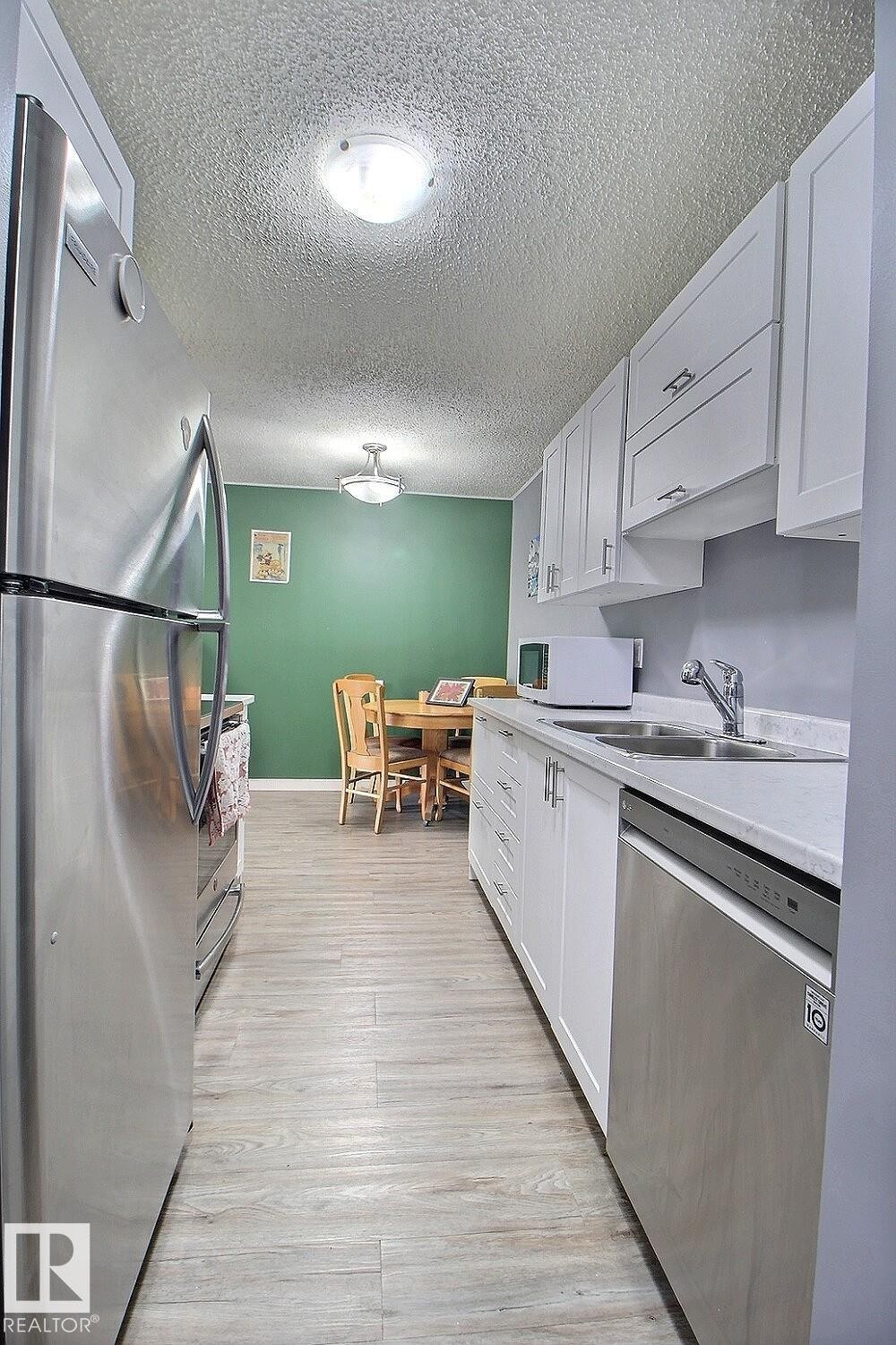 Galley kitchen featuring stainless steel appliances, white cabinetry, light countertops, and wood-finish flooring - #304 14004 26 St Nw, Edmonton, AB - Indoor Photo Showing Kitchen With Double Sink