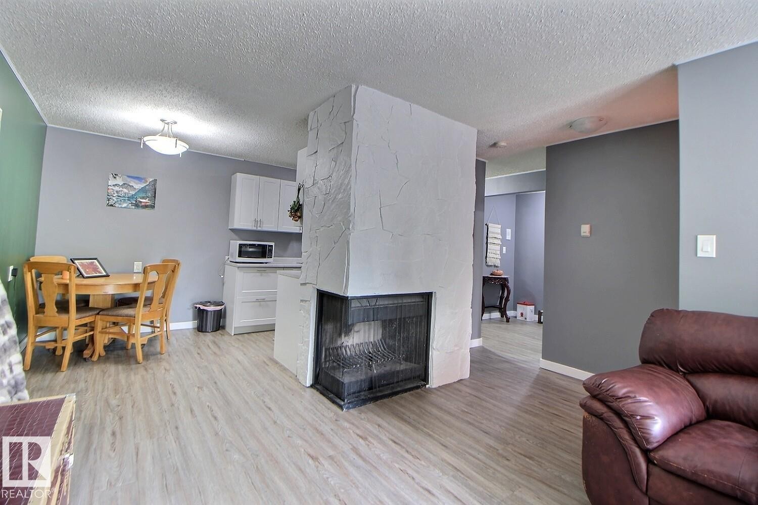 Central fireplace with a black firebox, light gray walls, wood-finish flooring, and white kitchen cabinetry - #304 14004 26 St Nw, Edmonton, AB - Indoor Photo Showing Living Room With Fireplace