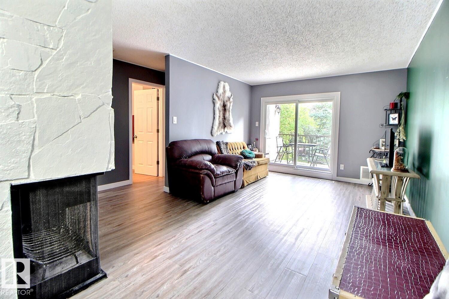 Light wood-finish flooring throughout, a stone-faced fireplace, and sliding glass doors leading to a balcony - #304 14004 26 St Nw, Edmonton, AB - Indoor Photo Showing Living Room