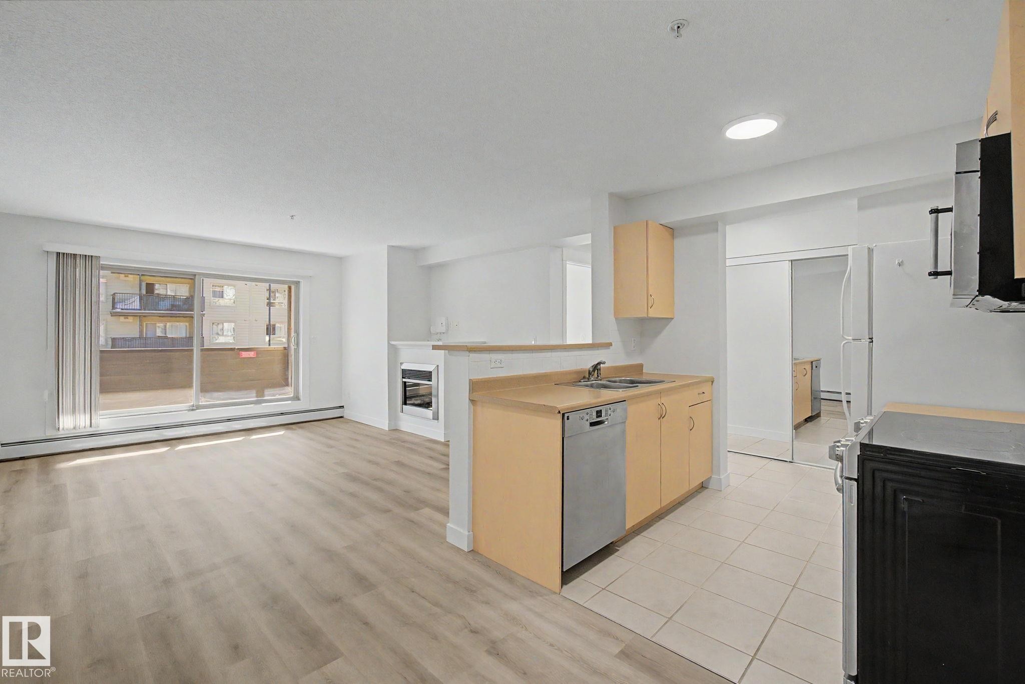 Open-concept living area with wood-finish flooring, sliding glass patio door, and integrated kitchen - #226 17011 67 Av Nw, Edmonton, AB - Outdoor With View