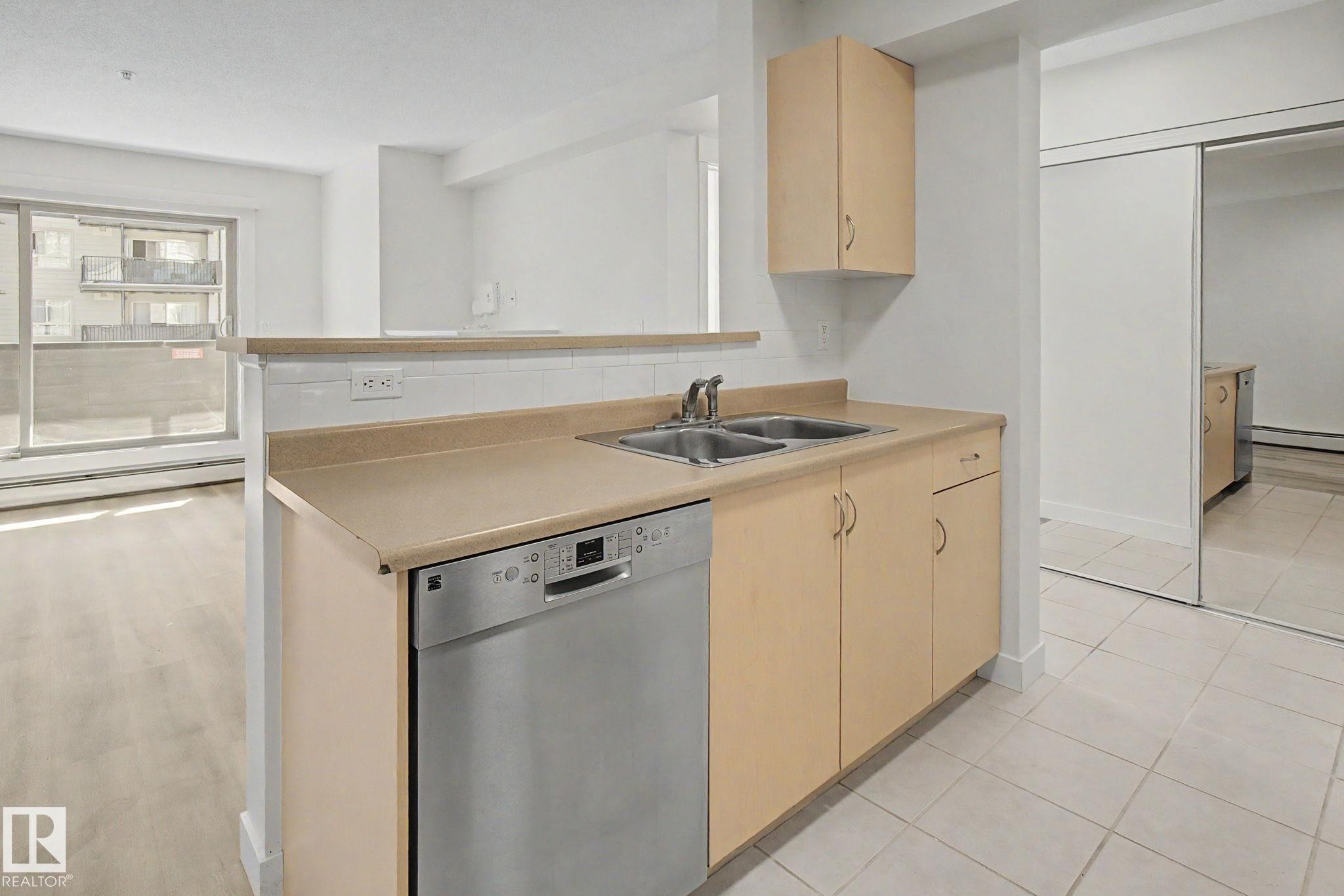 Kitchen featuring a stainless steel dishwasher, double basin sink, light wood-finish cabinetry, and a pass-through counter with a tiled backsplash - #226 17011 67 Av Nw, Edmonton, AB - Indoor Photo Showing Kitchen With Double Sink