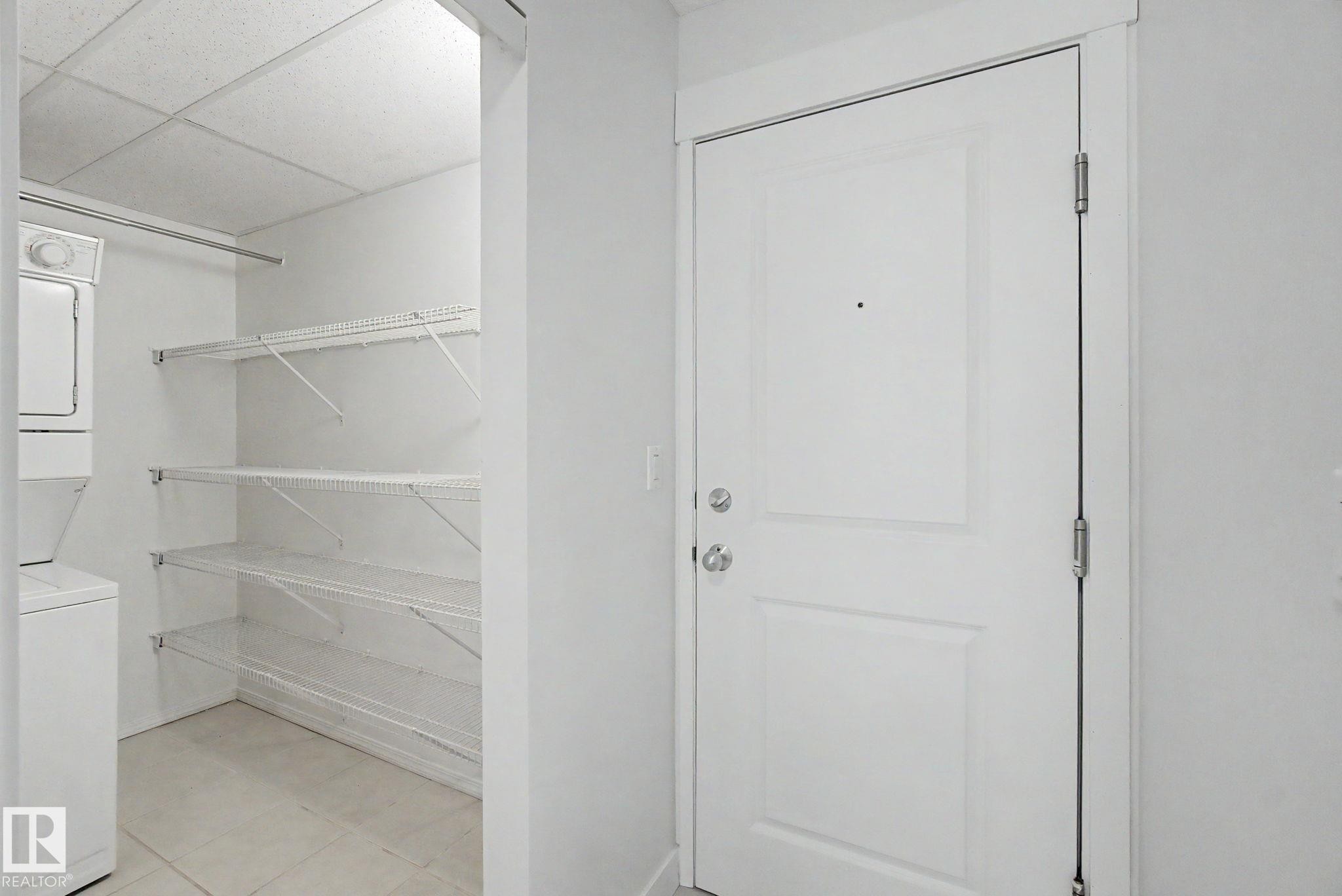 White paneled door with polished chrome hardware, adjacent to a utility closet featuring a stacked washer and dryer, wire shelving, and a hanging rod - #226 17011 67 Av Nw, Edmonton, AB - Indoor With Storage