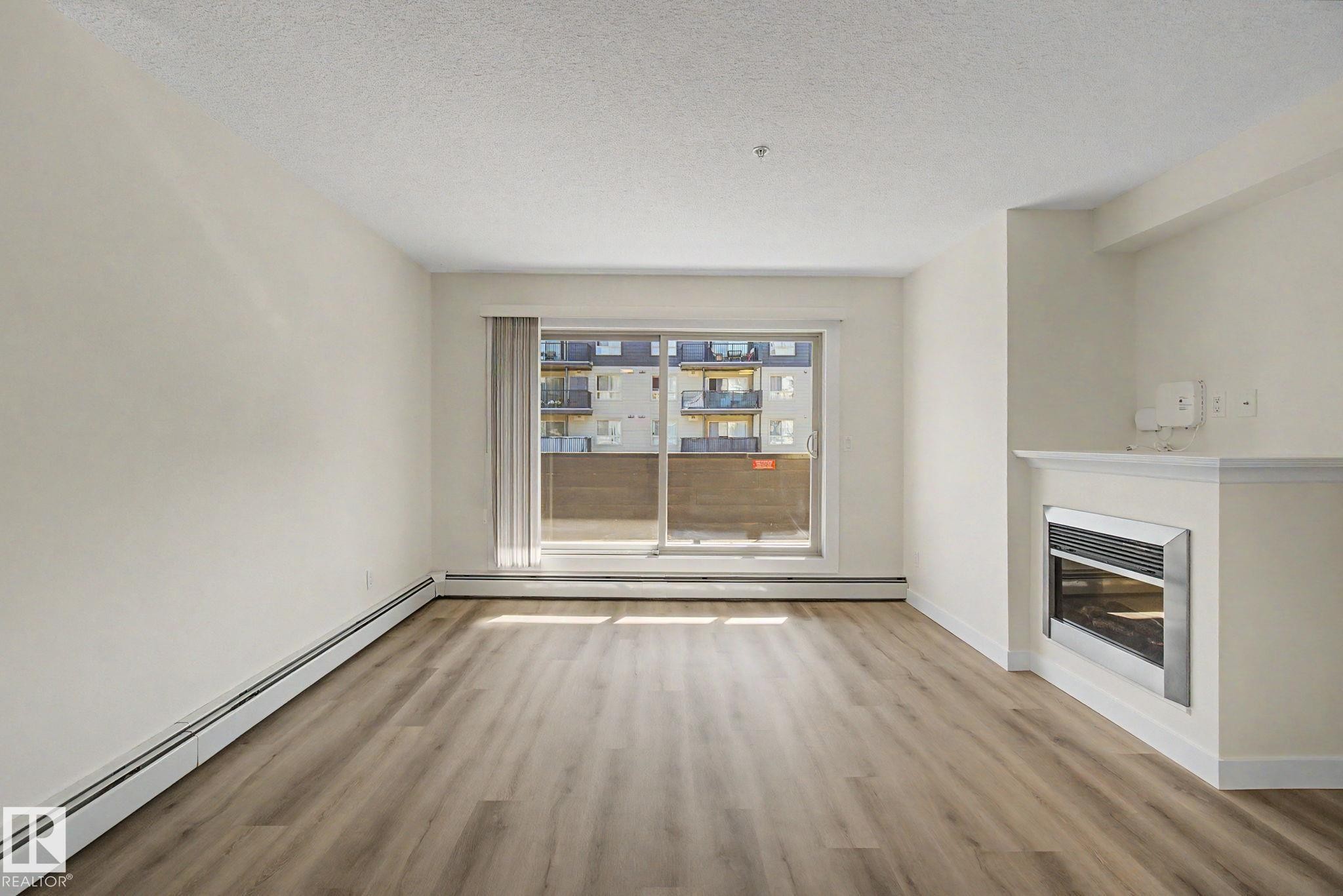 Spacious room featuring wood-finish flooring, a built-in fireplace with a mantel, and a sliding glass door - #226 17011 67 Av Nw, Edmonton, AB - Indoor With Fireplace
