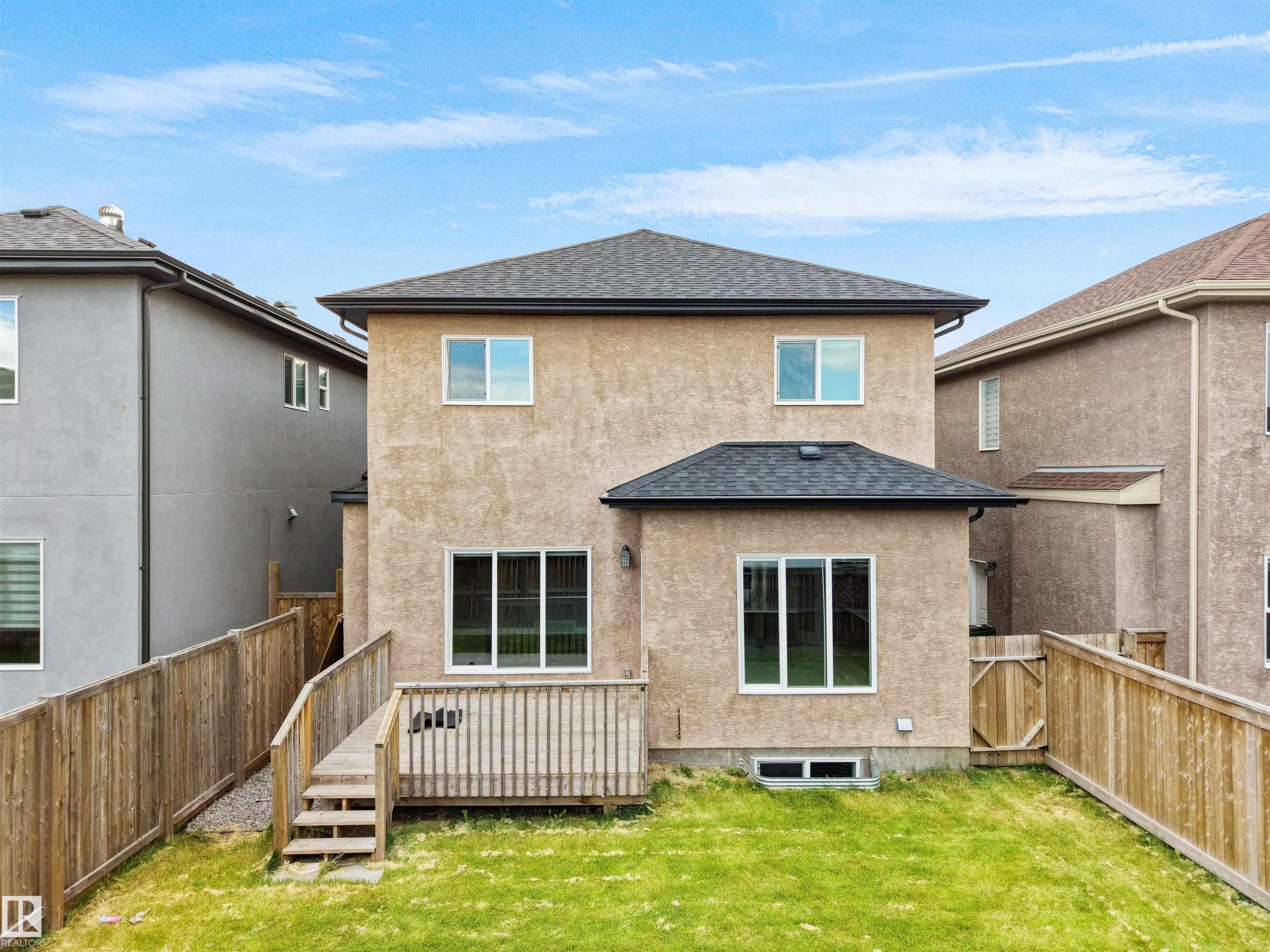 Rear exterior featuring stucco finish, a private wood fence, ground-level wood deck with steps, and multiple windows - 17020 62 Street Nw, Edmonton, AB - Outdoor With Exterior