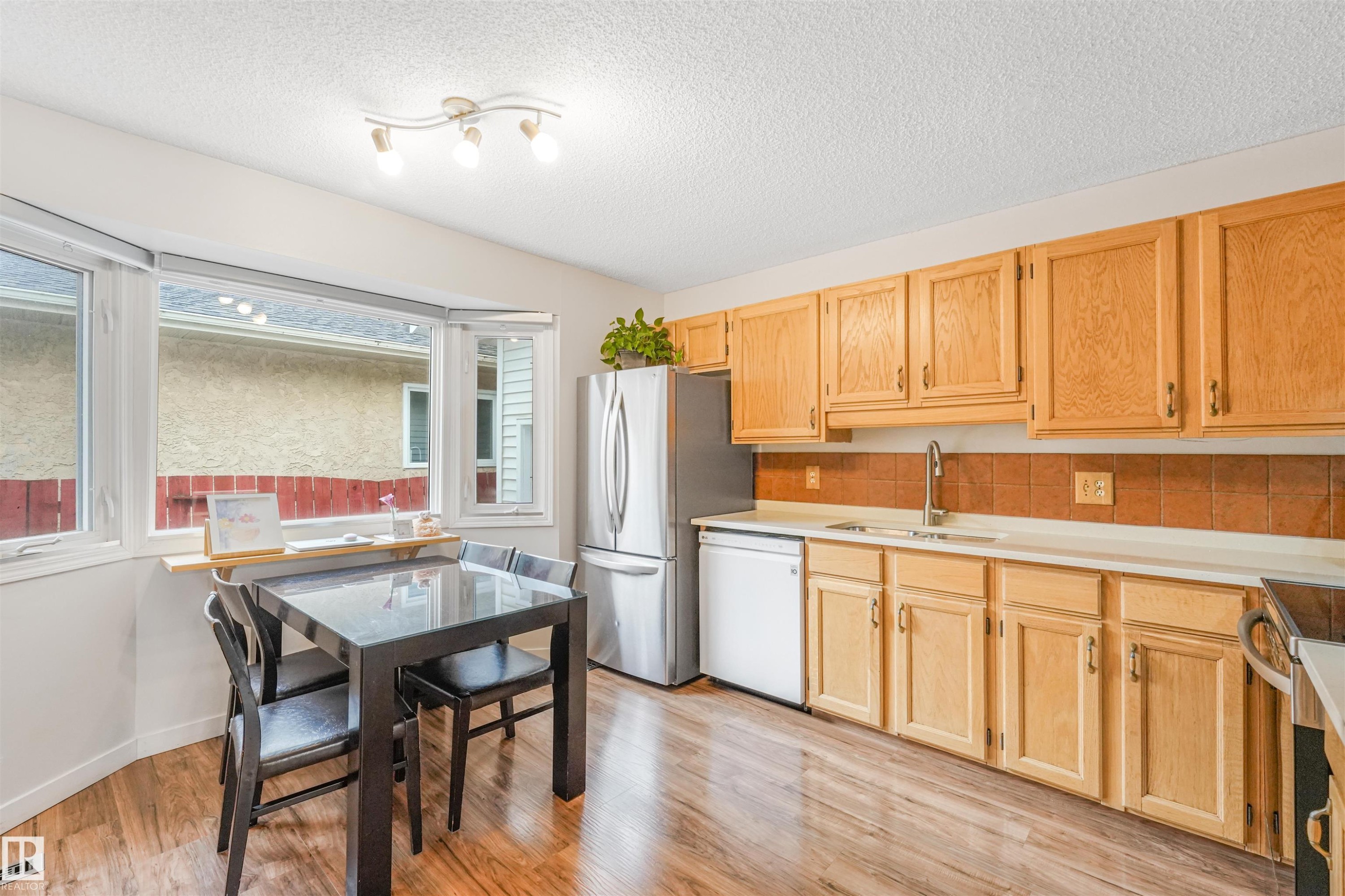 Kitchen featuring wood-finish flooring, natural wood cabinetry, a stainless steel refrigerator, and a bay window - 18611 57 Avenue Nw, Edmonton, AB - Indoor Photo Showing Kitchen