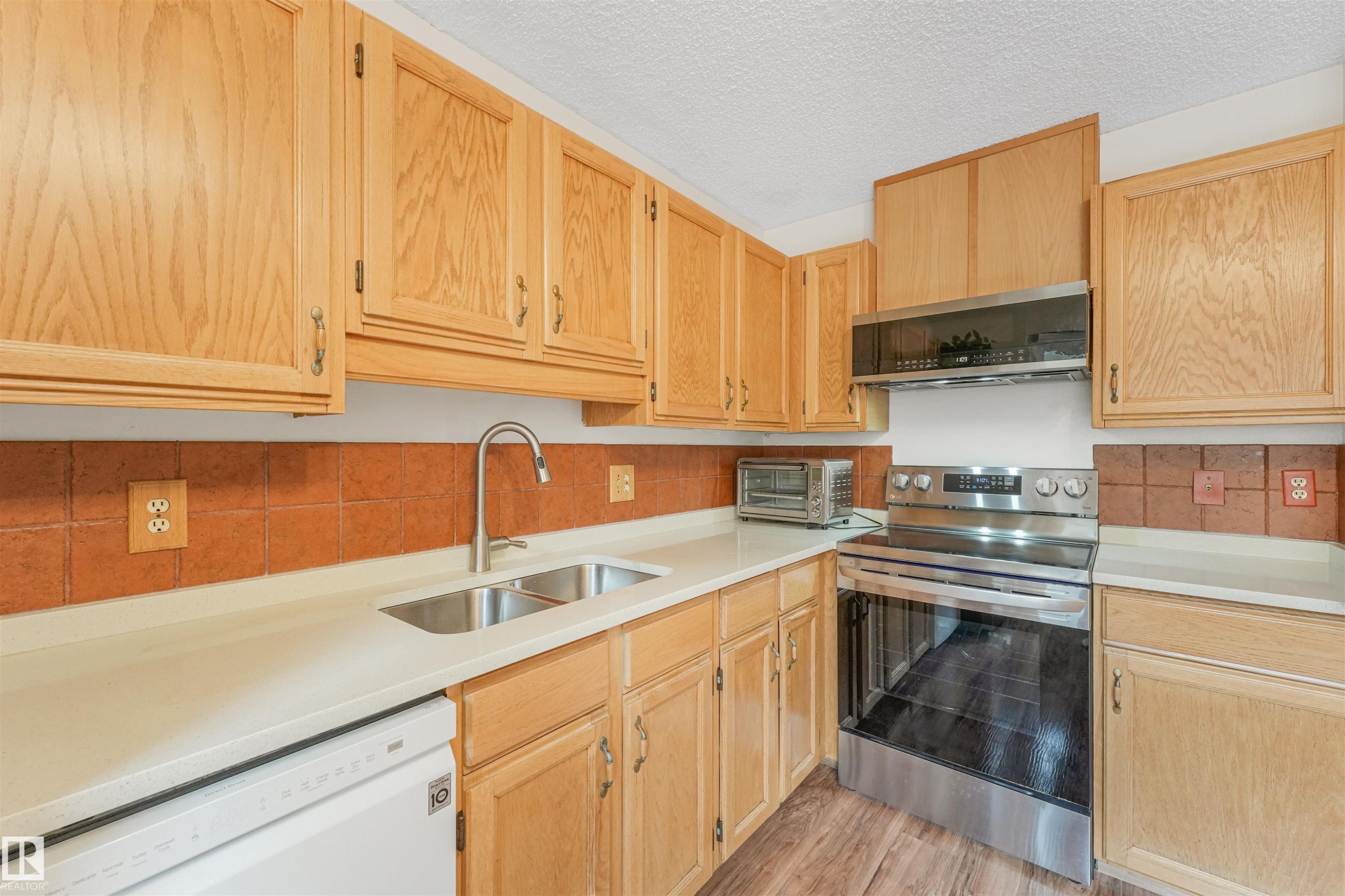 Kitchen featuring blonde wood cabinetry, stainless steel appliances, a double basin sink with a gooseneck faucet, light-toned countertops, and a terracotta tile backsplash - 18611 57 Avenue Nw, Edmonton, AB - Indoor Photo Showing Kitchen With Double Sink