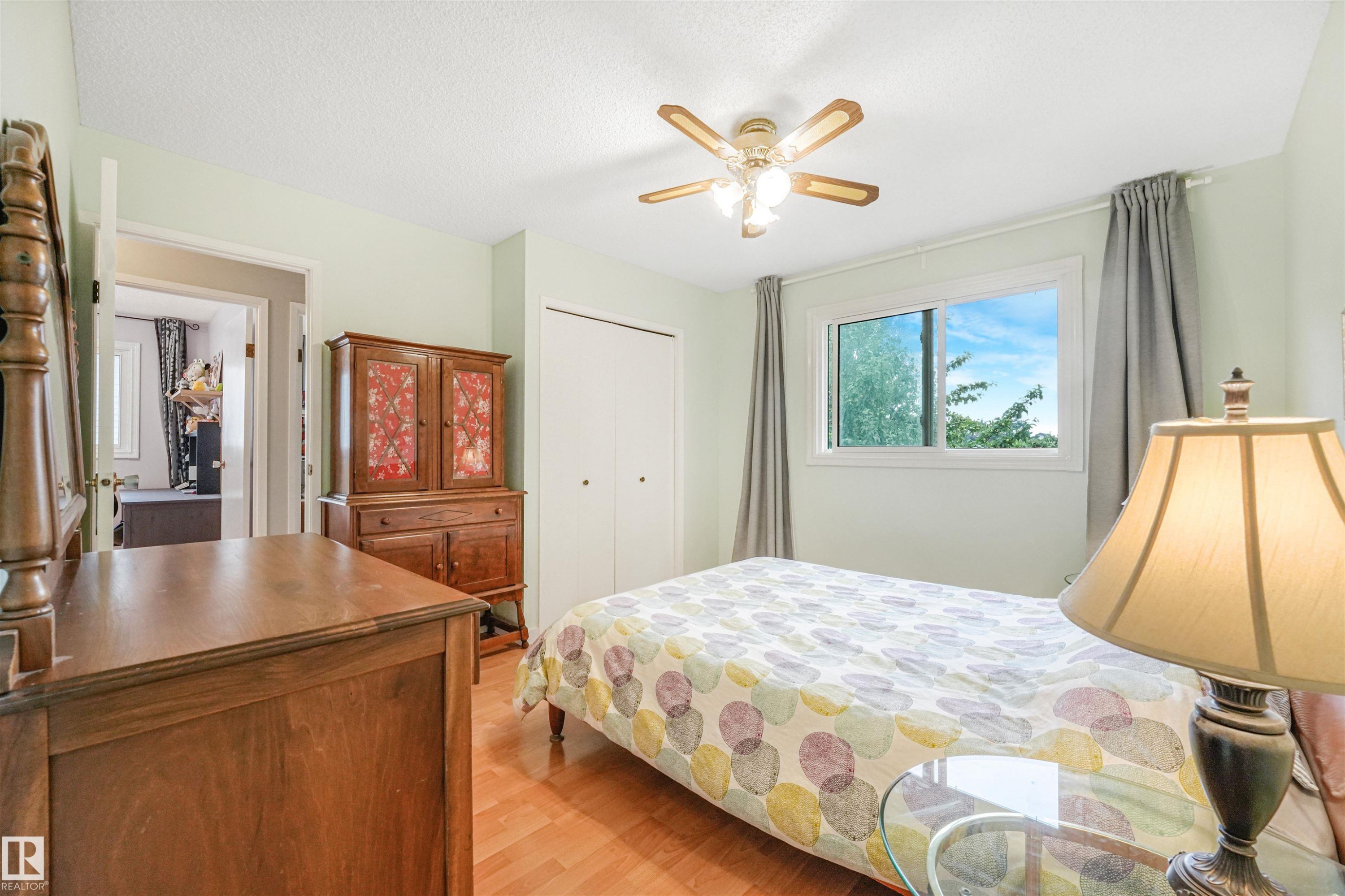 Light-filled room featuring wood-finish flooring, a window with exterior tree scenery, a ceiling fan with integrated lighting, and a closet with bifold doors - 18611 57 Avenue Nw, Edmonton, AB - Indoor Photo Showing Bedroom