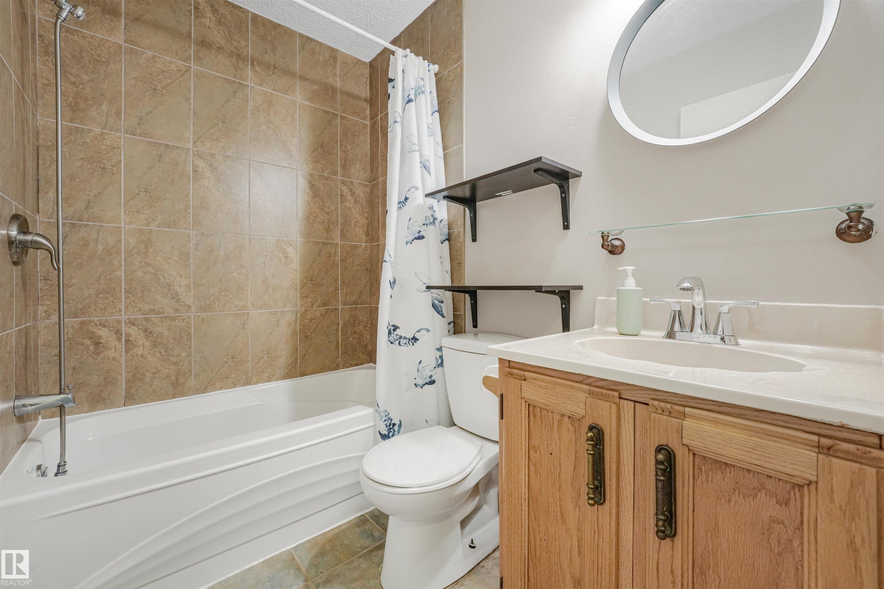 Bathroom featuring a tub/shower combination with tile surround, a single vanity with a light-colored countertop, and a circular wall mirror - 18611 57 Avenue Nw, Edmonton, AB - Indoor Photo Showing Bathroom