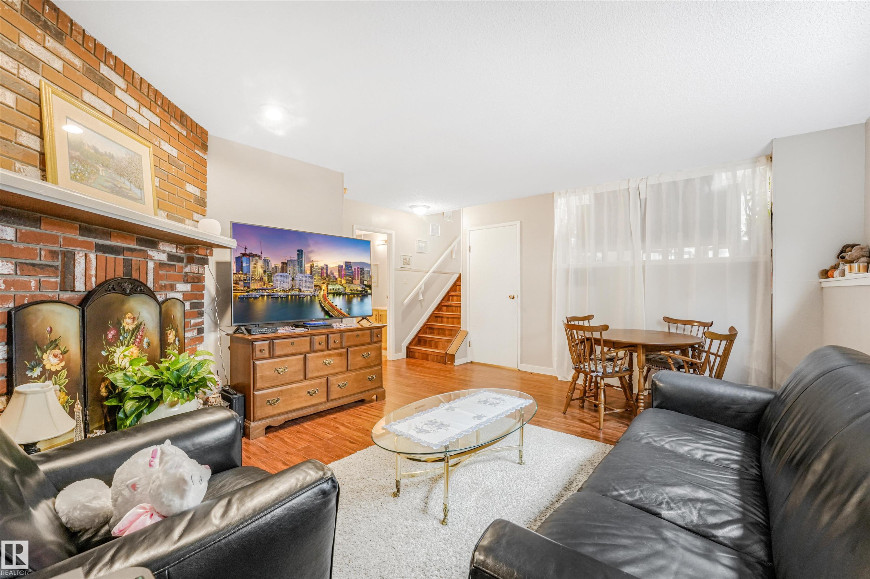 Living area featuring a brick fireplace, wood-finish flooring, staircase, and a dining alcove with a large window - 18611 57 Avenue Nw, Edmonton, AB - Indoor Photo Showing Living Room