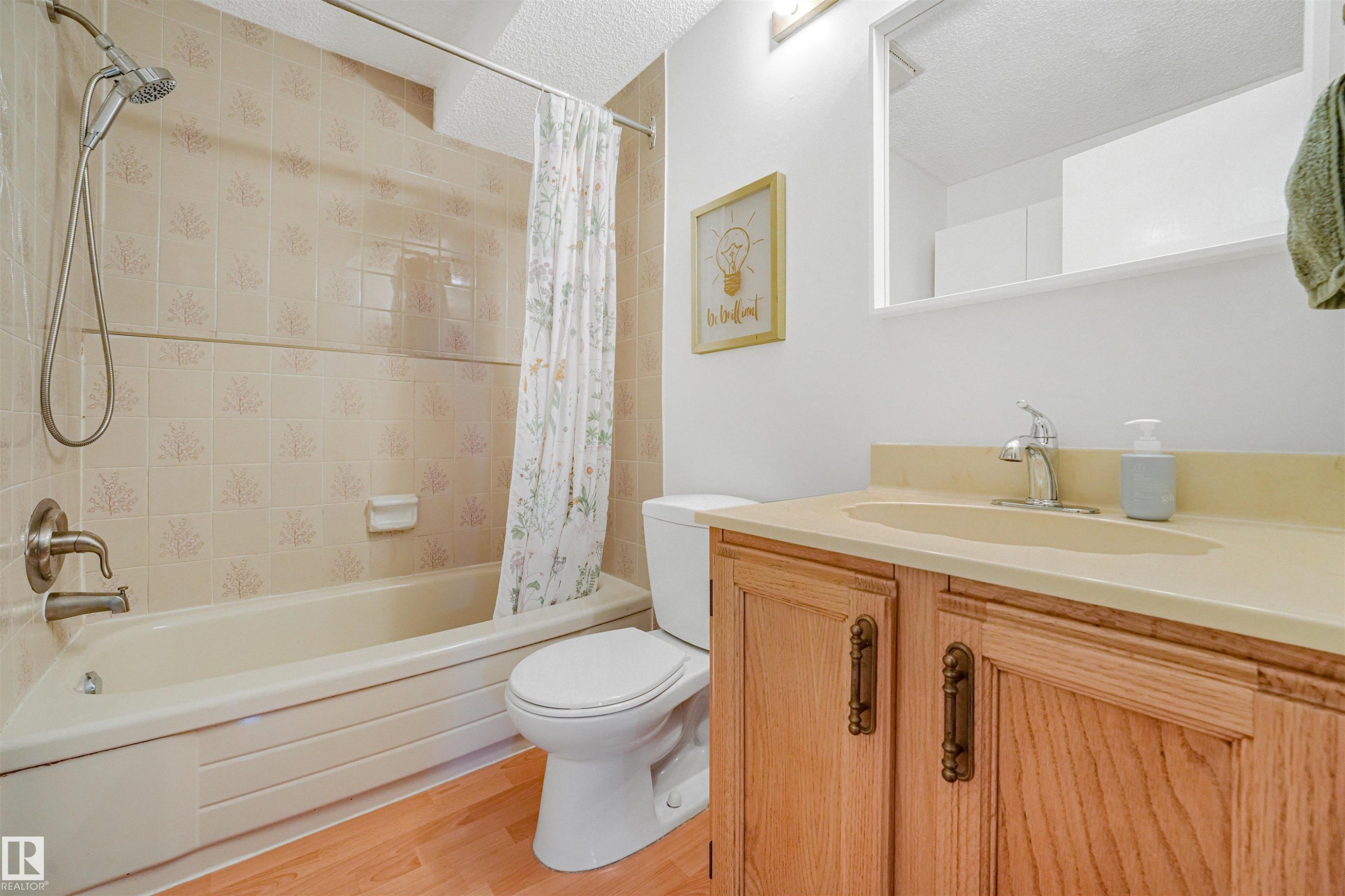 Bathroom featuring a light wood-finish vanity with a built-in sink, a rectangular mirror, a shower-over-tub combination with tiled surround, and wood-finish flooring - 18611 57 Avenue Nw, Edmonton, AB - Indoor Photo Showing Bathroom