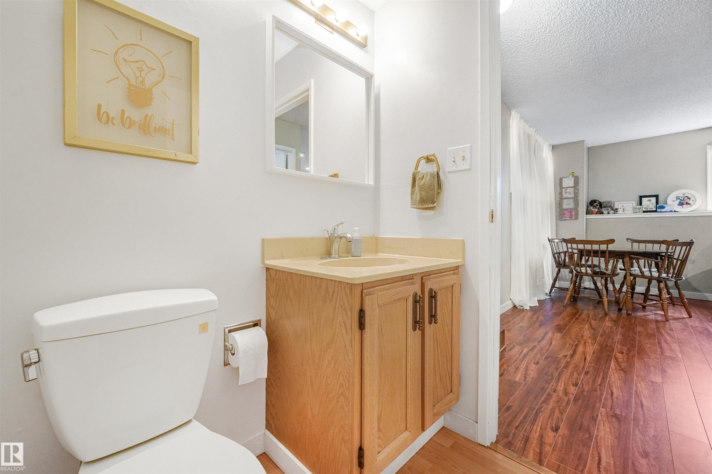 Bathroom featuring a white toilet, wood-finish vanity with a light-toned countertop, and an overhead mirror with strip lighting - 18611 57 Avenue Nw, Edmonton, AB - Indoor Photo Showing Bathroom