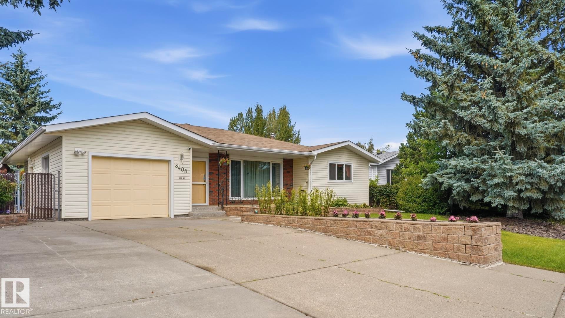 Exterior featuring light-colored siding, an attached garage, a concrete driveway, a stone retaining wall, and mature evergreen trees - 8408 176 Street Nw, Edmonton, AB - Outdoor