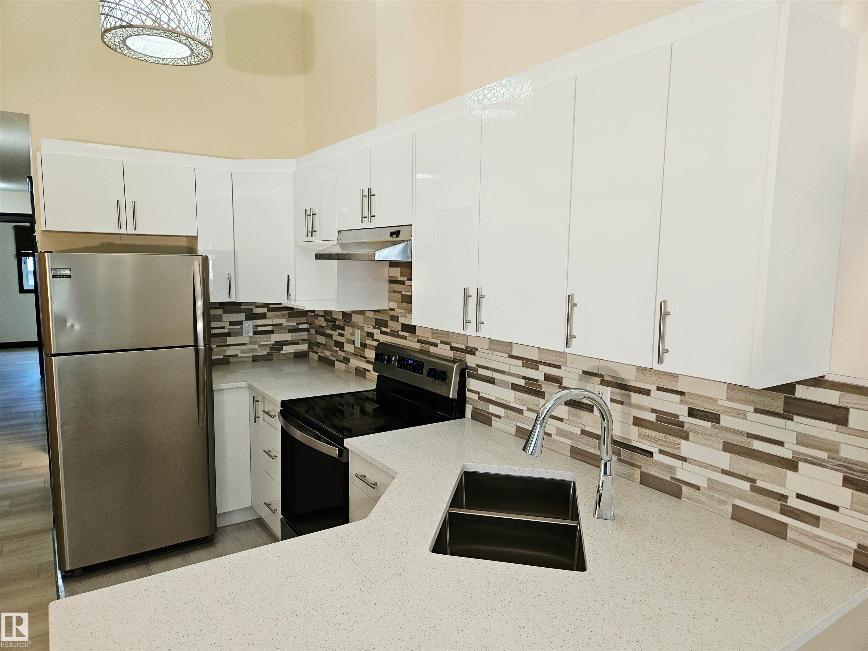 Galley kitchen featuring white cabinetry, light-toned countertops, a stainless steel double basin sink with a high-arch faucet, and a rectangular tile backsplash - 11625 88 Street Nw, Edmonton, AB