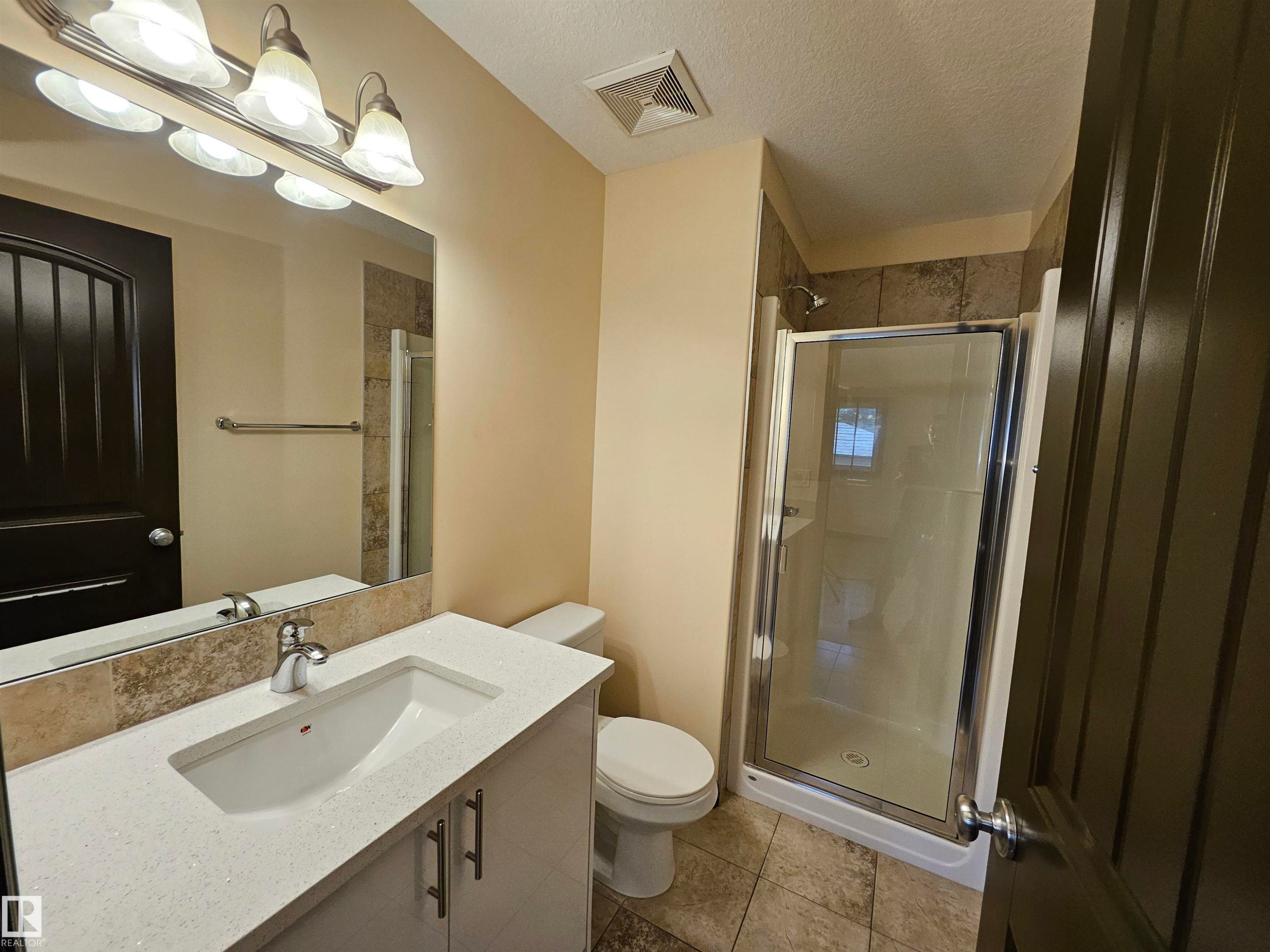 Vanity with an integrated sink and light-toned countertop, large mirror with overhead lighting, walk-in shower with glass enclosure, toilet, and tiled flooring - 11625 88 Street Nw, Edmonton, AB