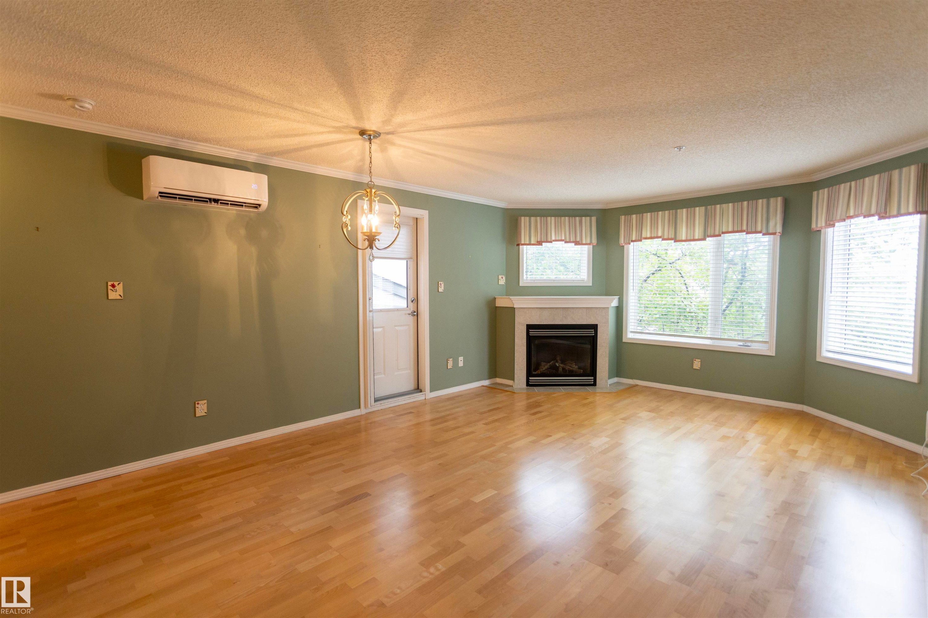 Hardwood-finish flooring, built-in fireplace with a light-toned mantel, multiple windows, wall-mounted air conditioning unit, and a decorative ceiling light fixture - #204 2420 108 St Nw, Edmonton, AB