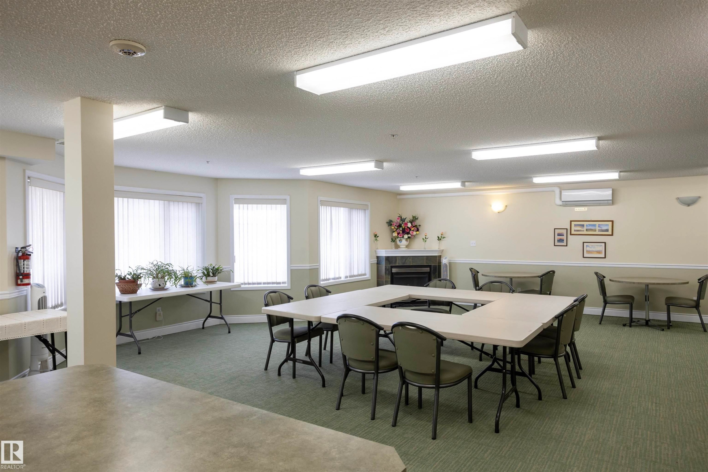 Spacious communal room with textured ceiling, multiple window units, wall-mounted air conditioning, and a built-in fireplace feature - #204 2420 108 St Nw, Edmonton, AB
