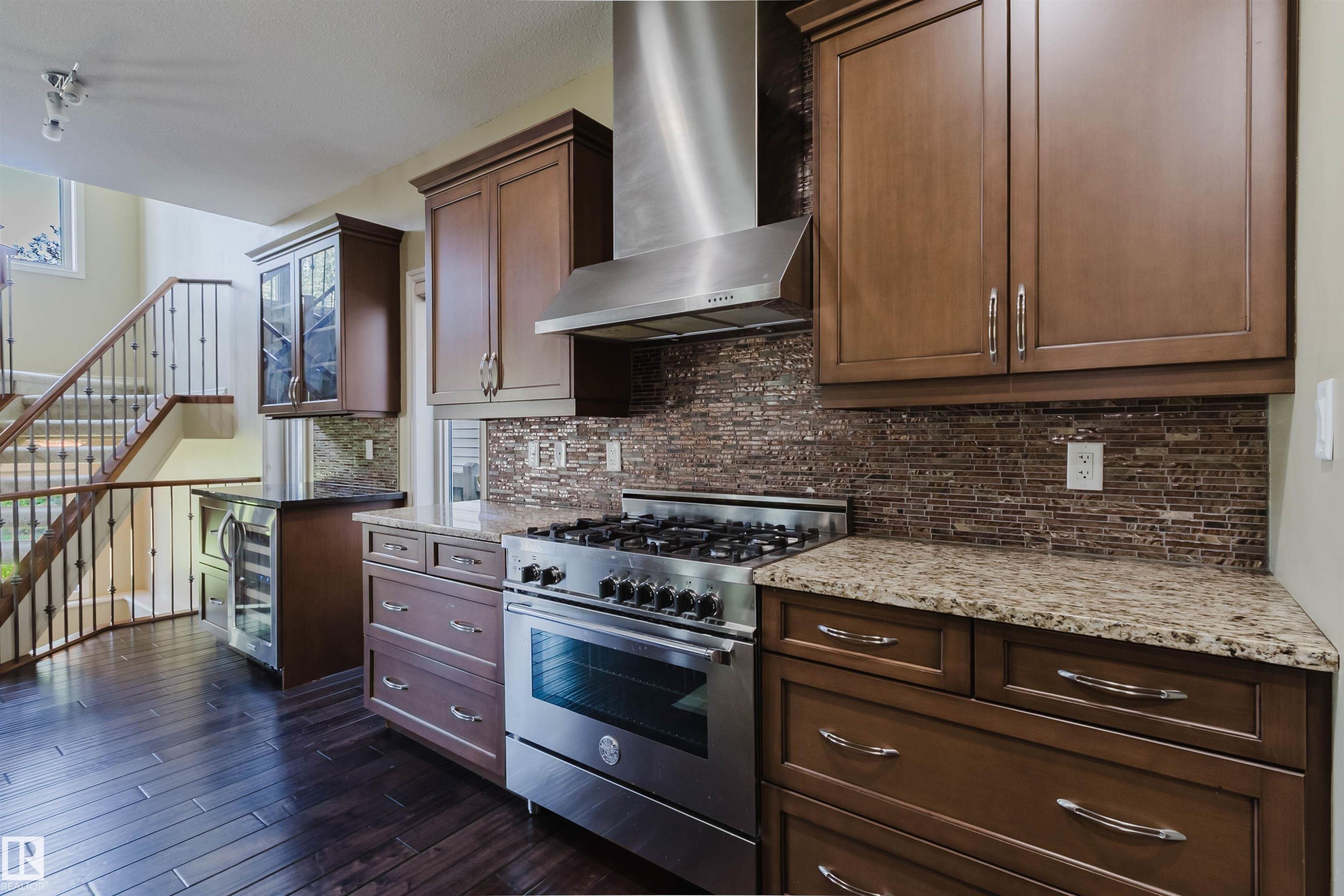 Stainless steel gas range with a matching range hood, mosaic tile backsplash, wood-finish flooring, and a built-in wine refrigerator - 864 Armitage Wynd Sw, Edmonton, AB - Indoor Photo Showing Kitchen