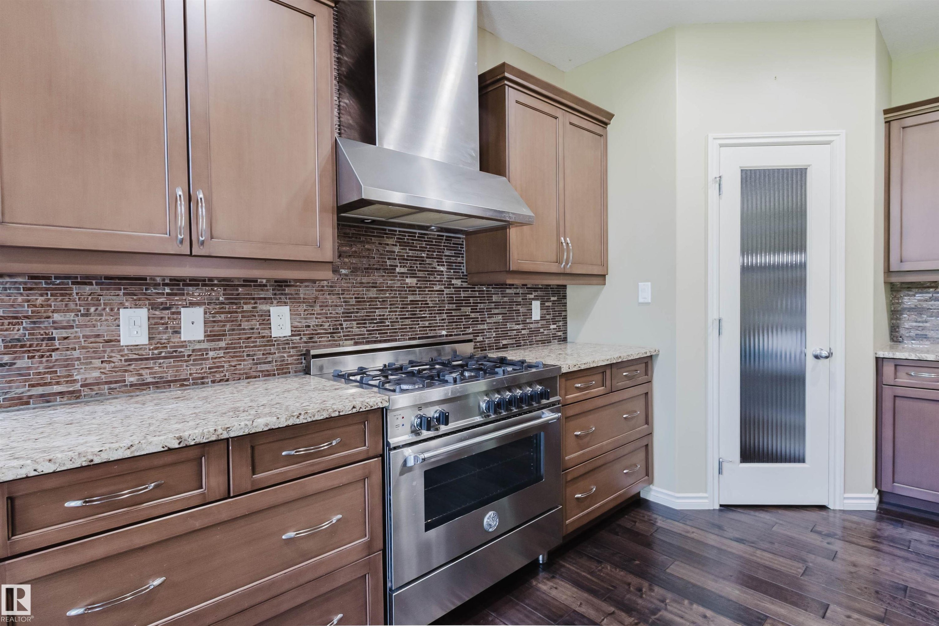 Stainless steel gas range with coordinating range hood, brown wood-finish cabinetry, granite countertops, and a full-height tile backsplash - 864 Armitage Wynd Sw, Edmonton, AB - Indoor Photo Showing Kitchen With Upgraded Kitchen
