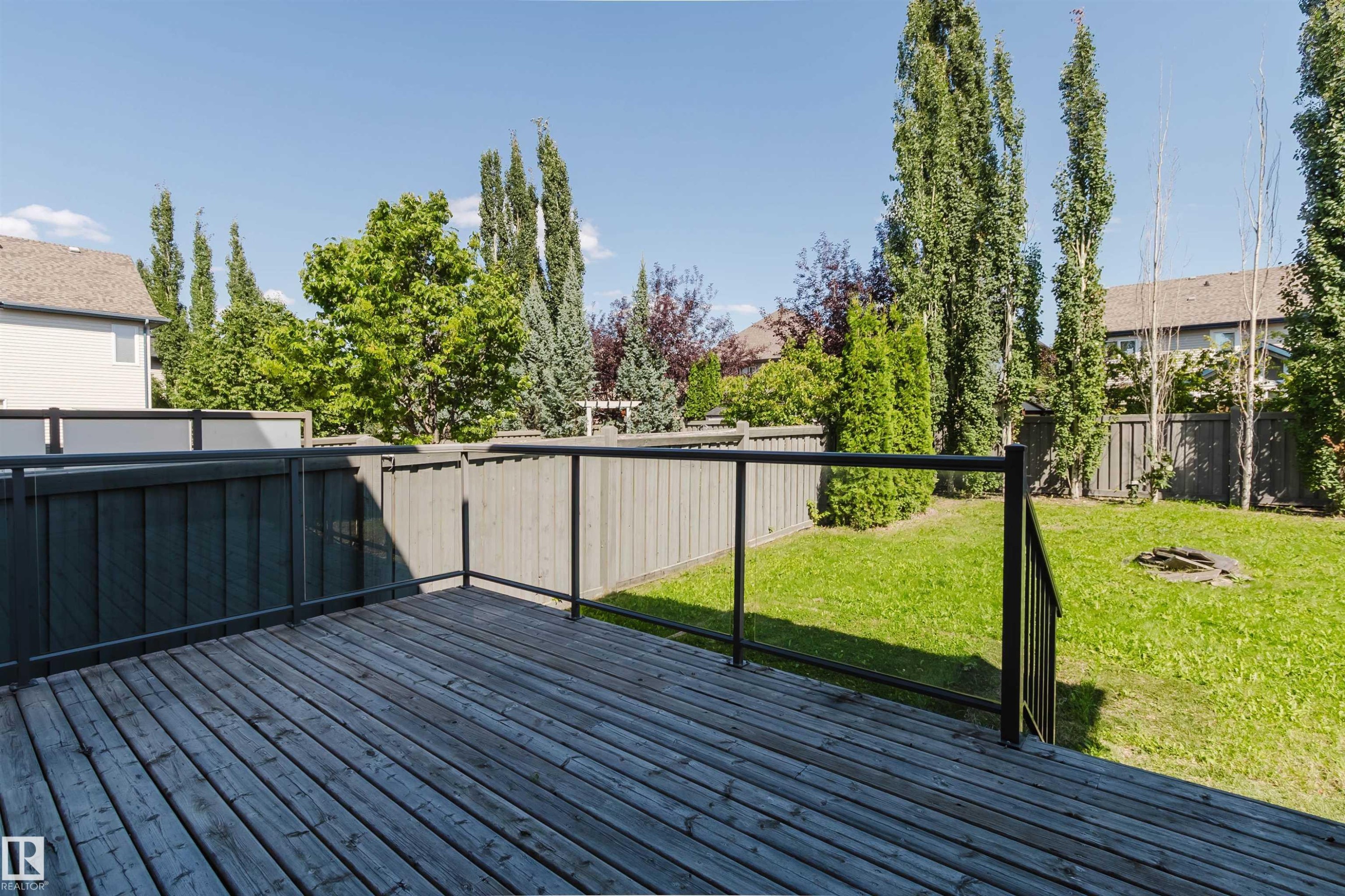 Expansive wood-finish deck with black metal railings, overlooking a fenced yard featuring mature trees and a grass lawn - 864 Armitage Wynd Sw, Edmonton, AB - Outdoor