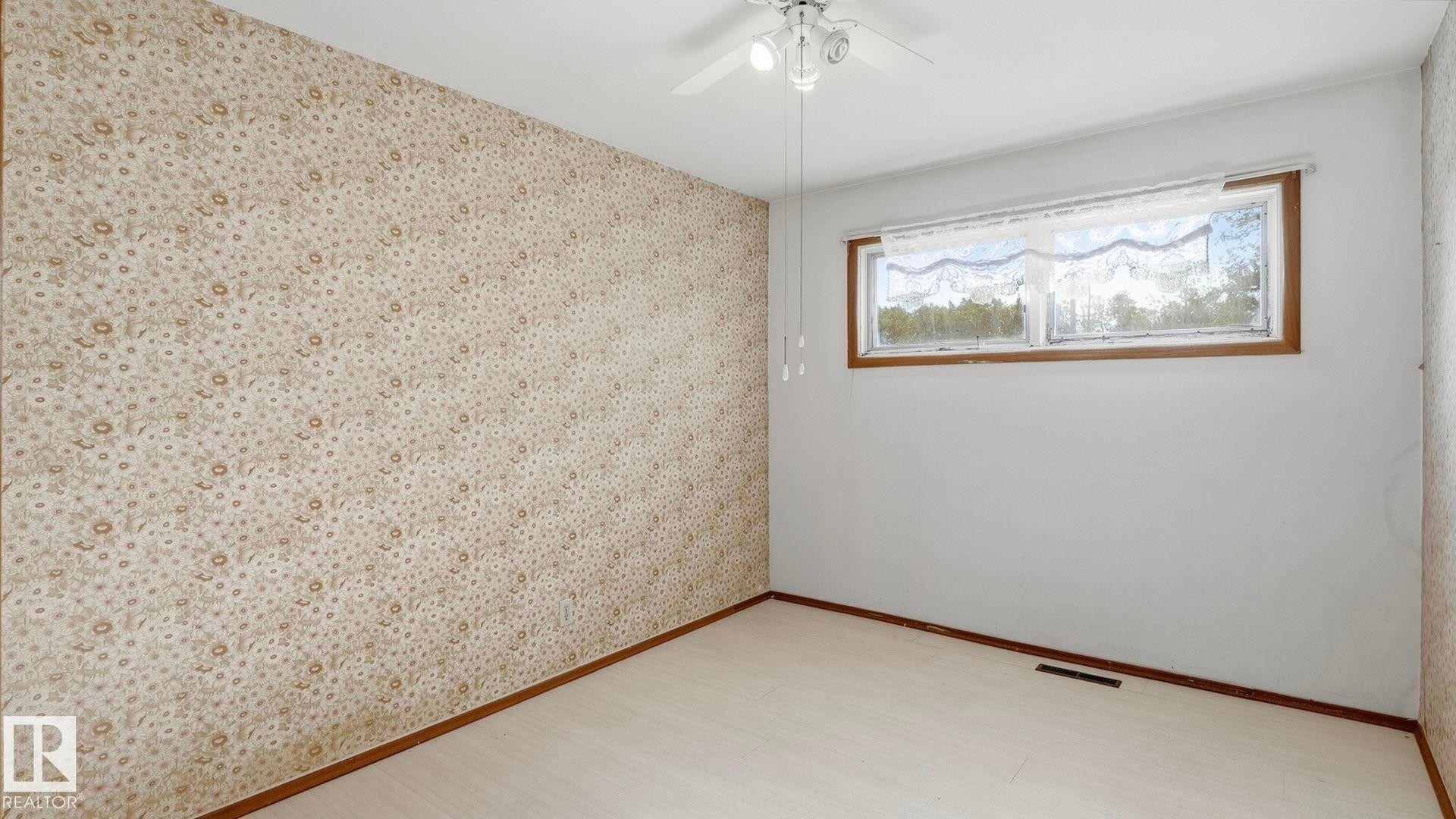 Patterned wallpaper accent wall, wood-finish flooring, and a ceiling fan with integrated lighting - 52012 Hwy 21, Rural Strathcona County, AB - Indoor Photo Showing Other Room