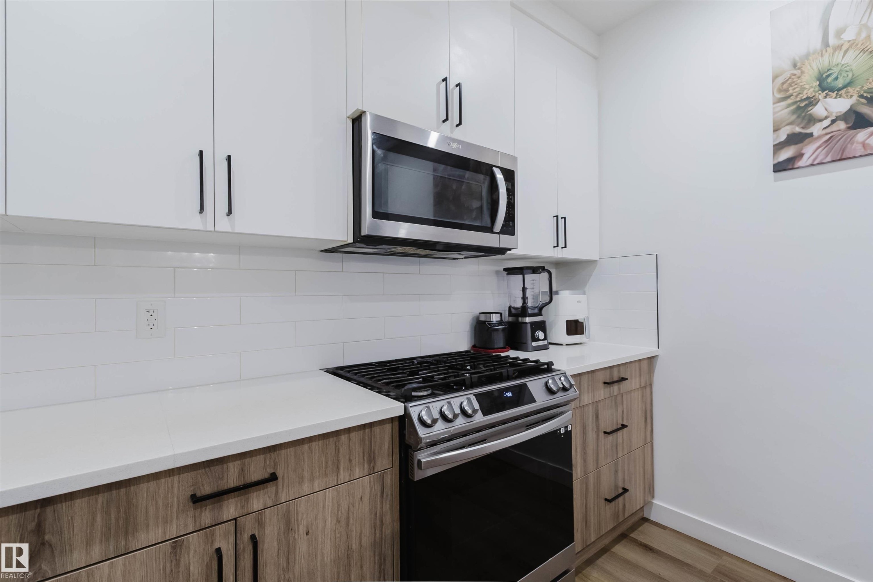 White upper cabinetry with black hardware, wood-finish lower cabinetry, white subway tile backsplash, white countertops, and stainless steel microwave and range - 104 Meadowview Way, Leduc, AB - Indoor Photo Showing Kitchen With Upgraded Kitchen