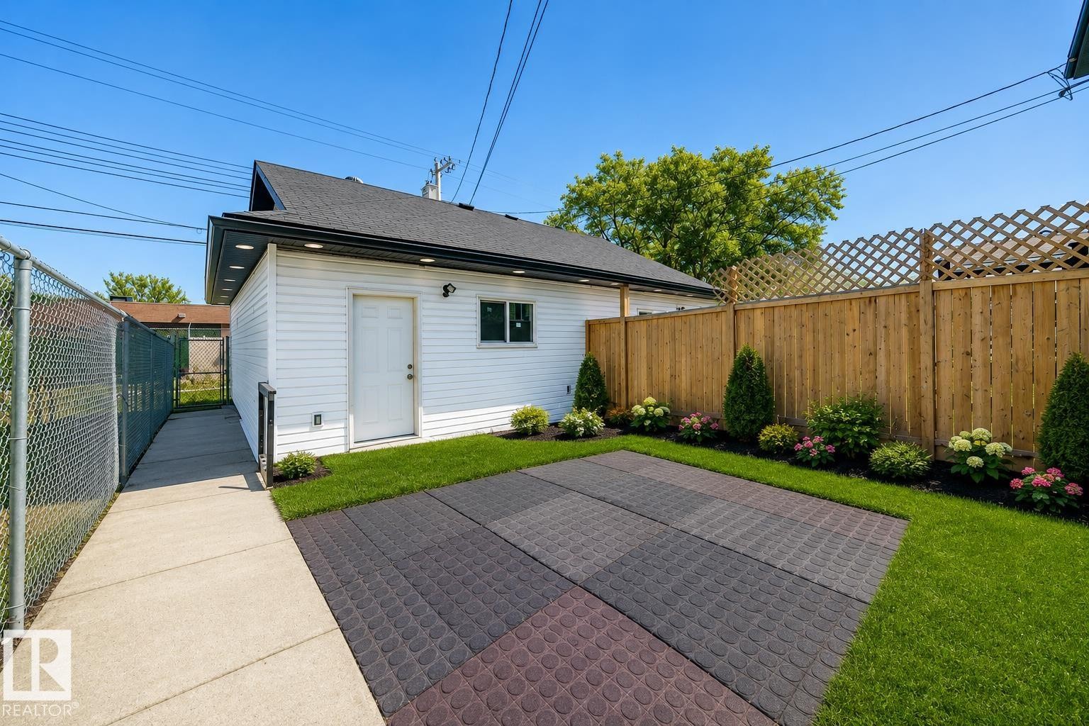 Virtually enhanced image of Paved patio area, manicured lawn, wood privacy fence with lattice accent, landscaped garden beds, and an exterior building with white siding - 11727 83 Street Nw, Edmonton, AB - Outdoor With Exterior