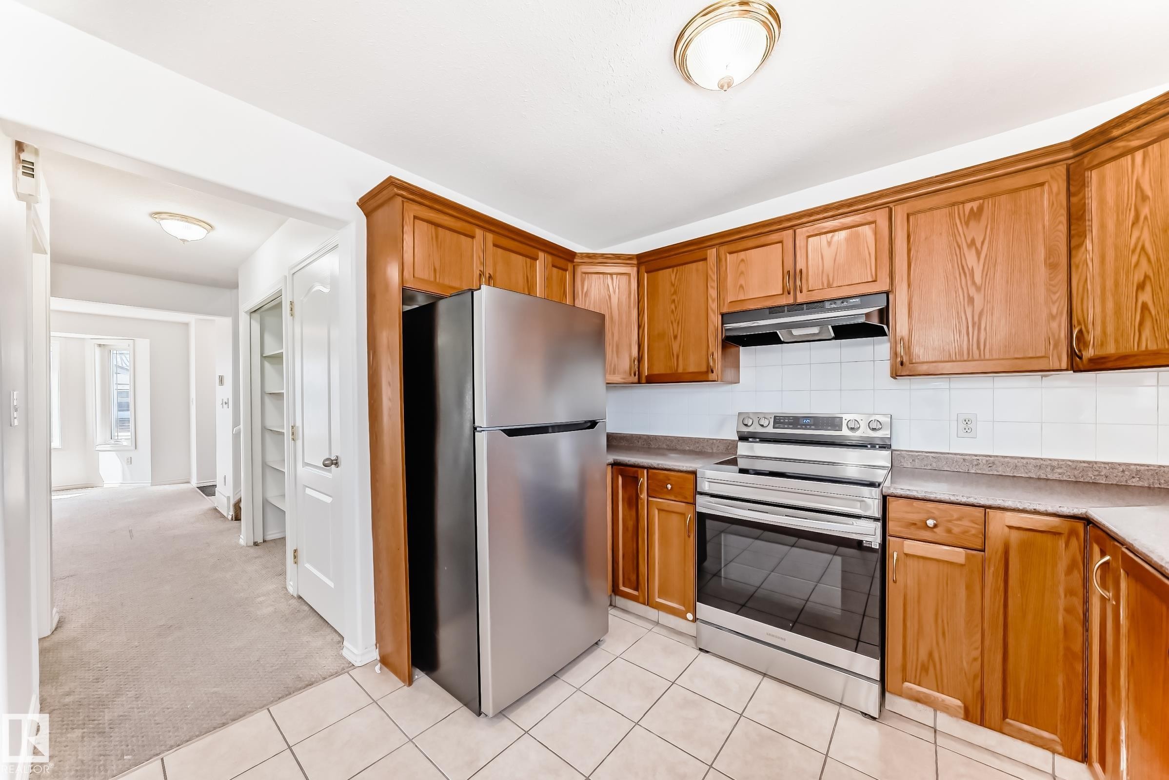 Kitchen featuring wood cabinetry, stainless steel appliances, white tile backsplash, light-toned countertops, and tile flooring - 11727 83 Street Nw, Edmonton, AB - Indoor Photo Showing Kitchen