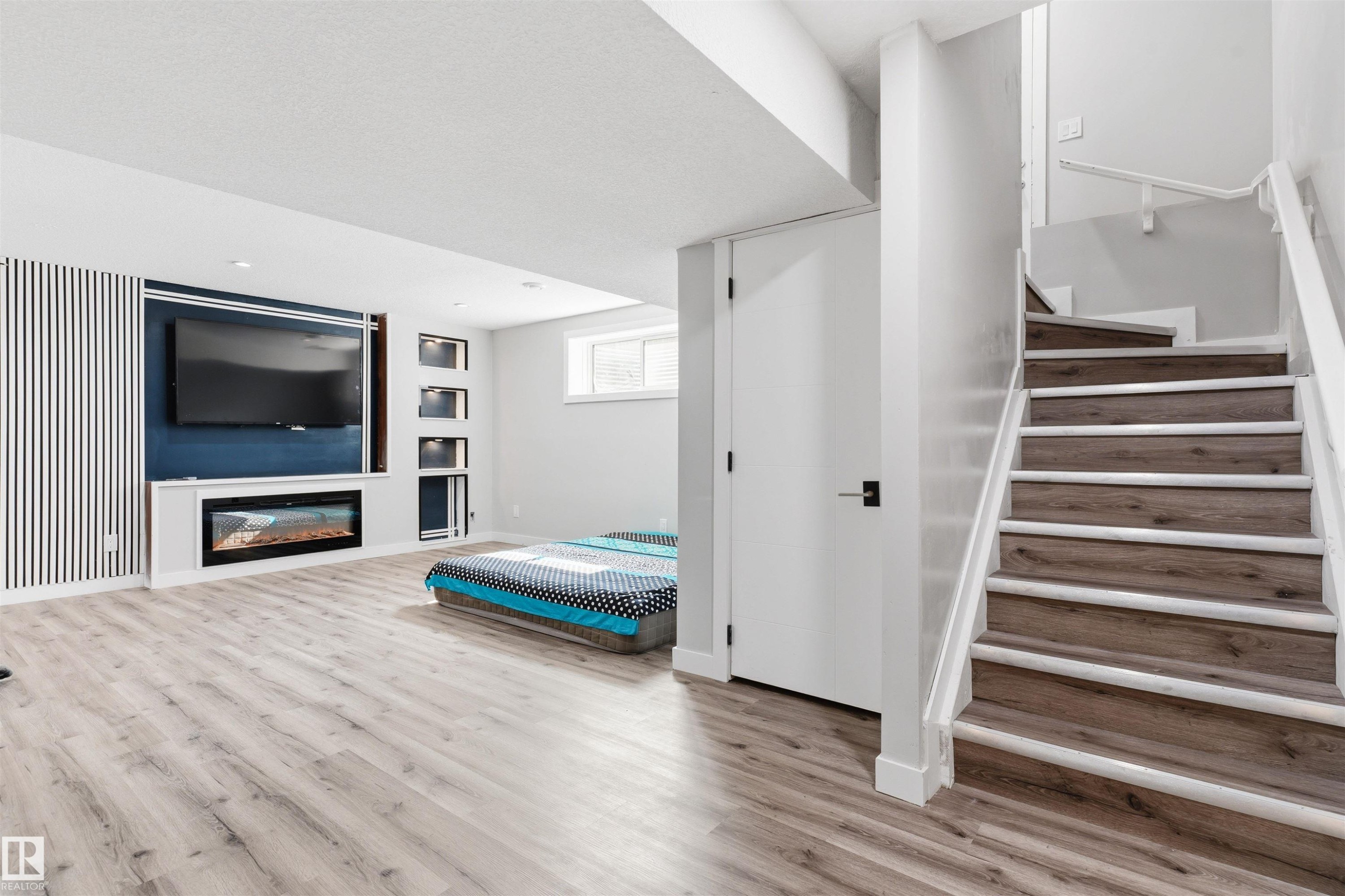 Recessed media wall with electric fireplace and integrated shelving, light wood-finish flooring, white painted staircase with wood-finish treads, white interior door with matte black hardware, and an egress window - 1103 26 Avenue Nw, Edmonton, AB