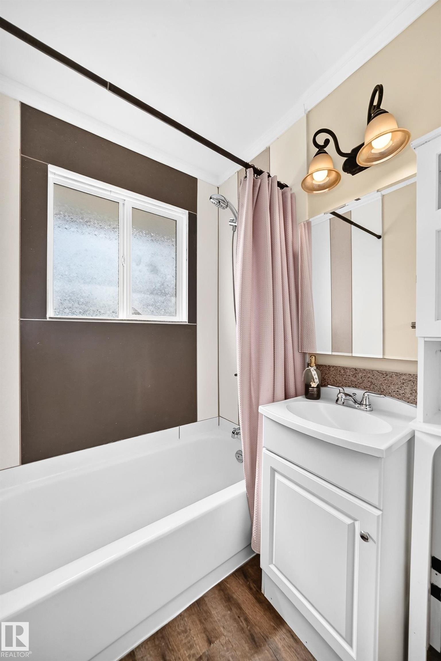 White bathtub with shower, white vanity cabinet with sink, decorative wall sconce lighting, and wood-finish flooring - 50 Rocque, Fort Saskatchewan, AB - Indoor Photo Showing Bathroom