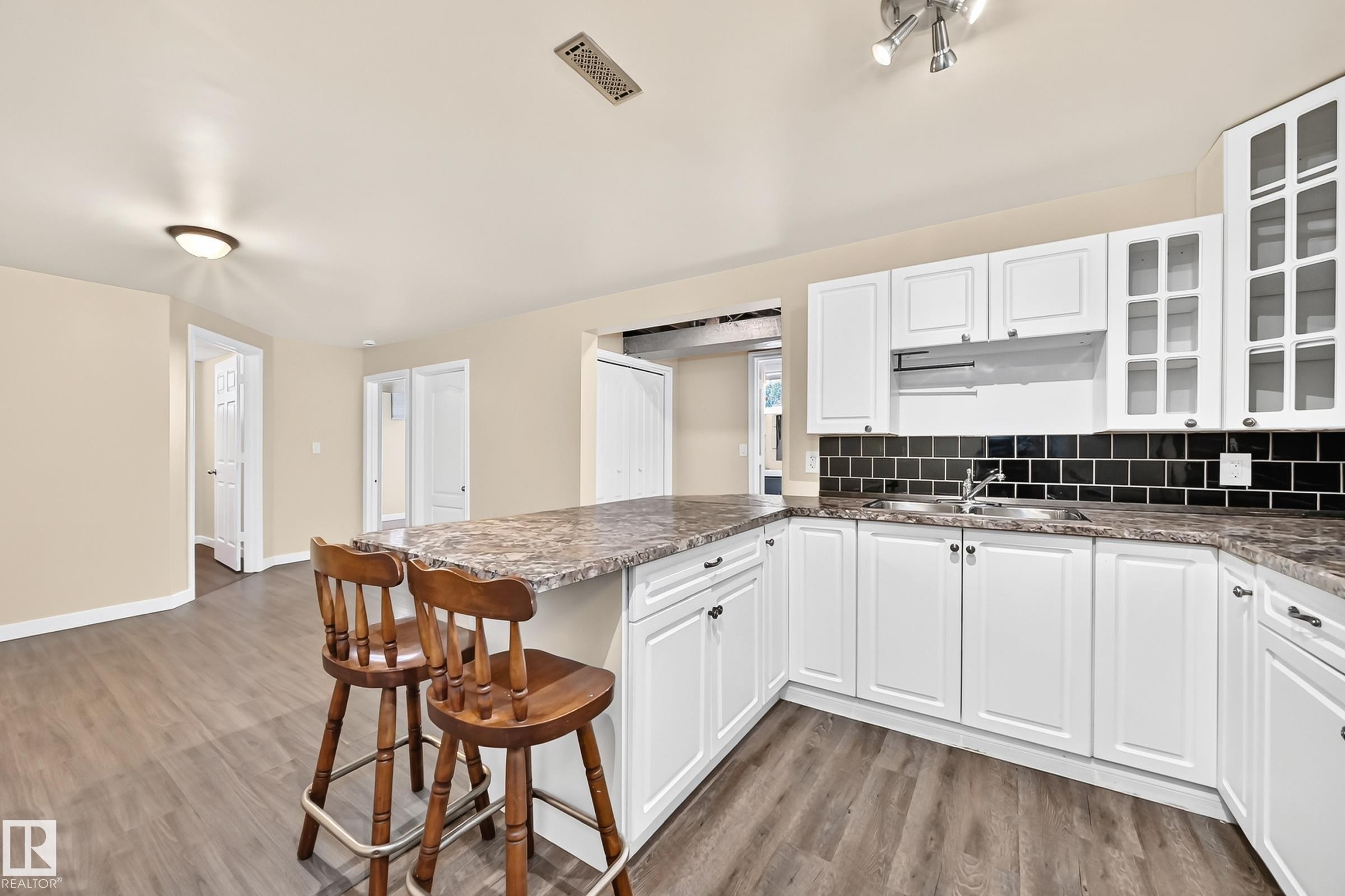 Wood-finish flooring throughout, white cabinetry with glass-front accents, dark subway tile backsplash, and a built-in kitchen island with stone-look countertop - 50 Rocque, Fort Saskatchewan, AB - Indoor Photo Showing Kitchen