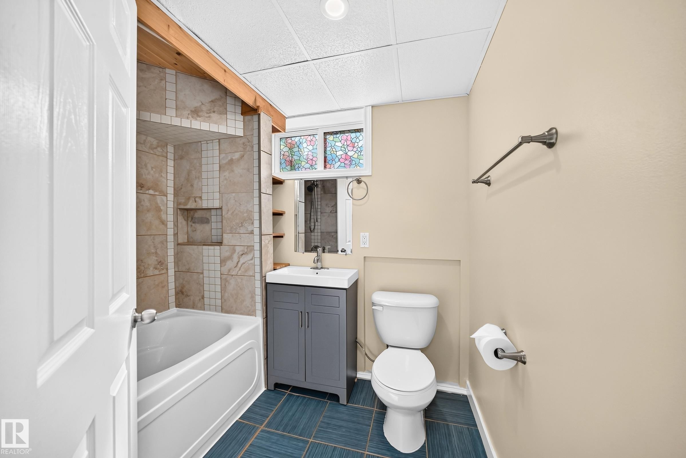 Ceramic tile flooring, tiled bathtub surround with built-in niche, white bathtub, vanity with integrated sink, and stained-glass window - 50 Rocque, Fort Saskatchewan, AB - Indoor Photo Showing Bathroom