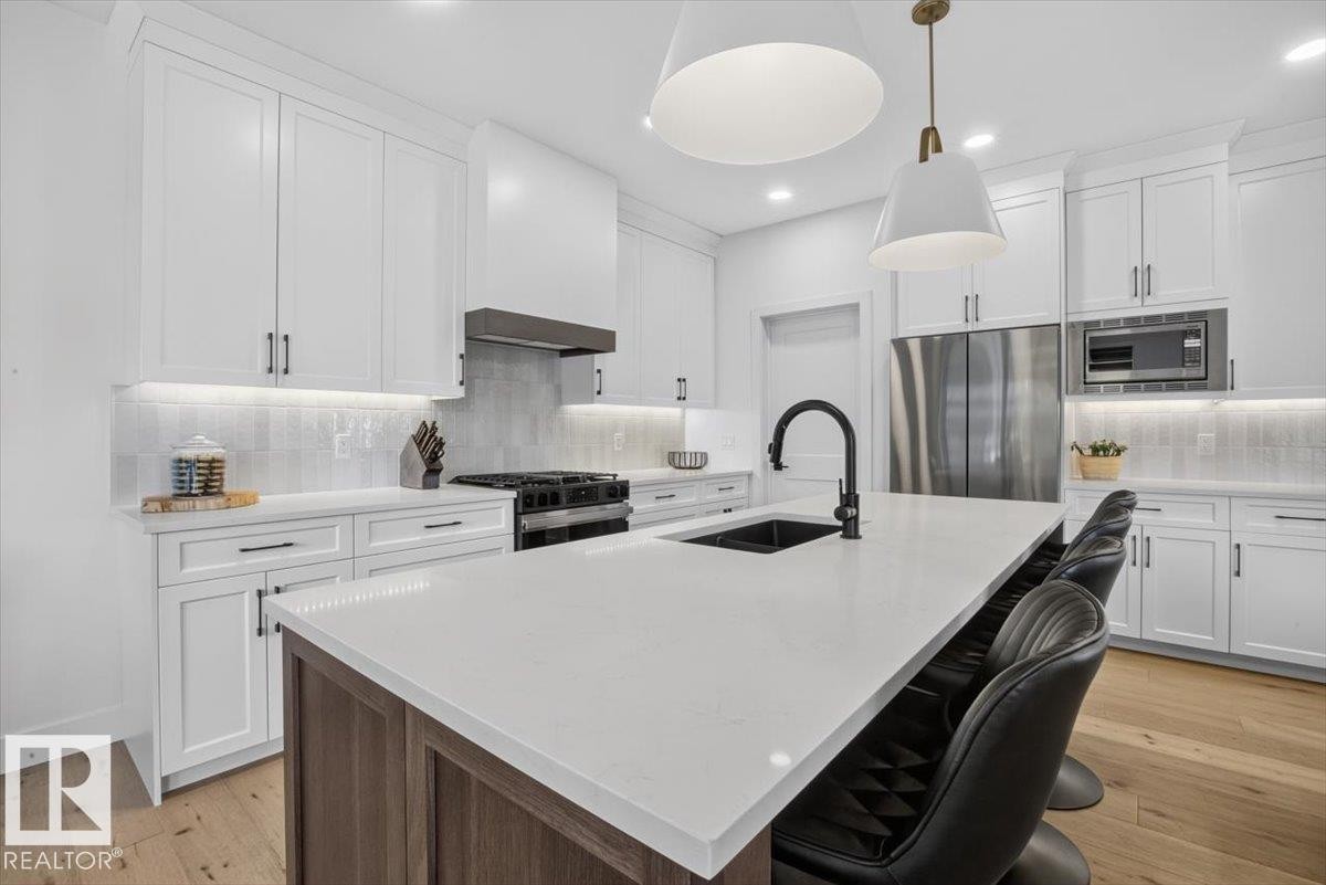 Kitchen featuring a spacious island with counter seating, stainless steel appliances, white shaker cabinetry, tiled backsplash, and wood-finish flooring - 94 Harvest Crescent, Ardrossan, AB - Indoor Photo Showing Kitchen With Stainless Steel Kitchen With Upgraded Kitchen
