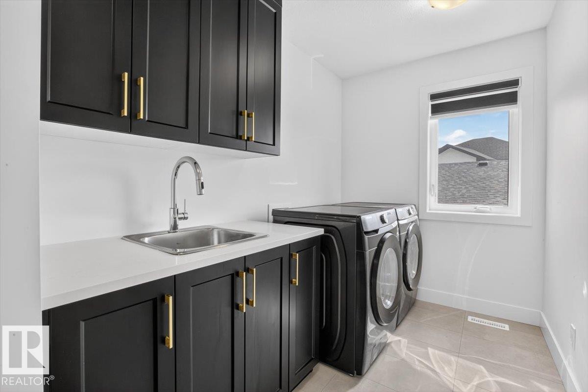 Black shaker-style cabinetry with gold hardware, white countertop, stainless steel sink with gooseneck faucet, and light-toned floor tile - 94 Harvest Crescent, Ardrossan, AB - Indoor Photo Showing Laundry Room