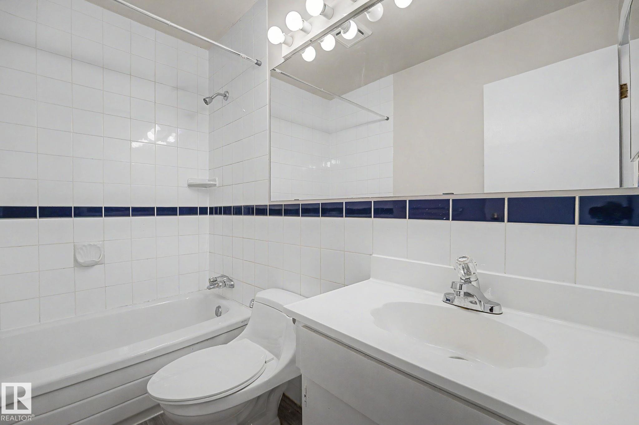 Bathroom featuring a white tile shower surround with a dark blue accent band, a white vanity with an integrated sink, a chrome faucet, a wall-mounted mirror, and a vanity light fixture - 1096 Knottwood Road Nw, Edmonton, AB - Indoor Photo Showing Bathroom