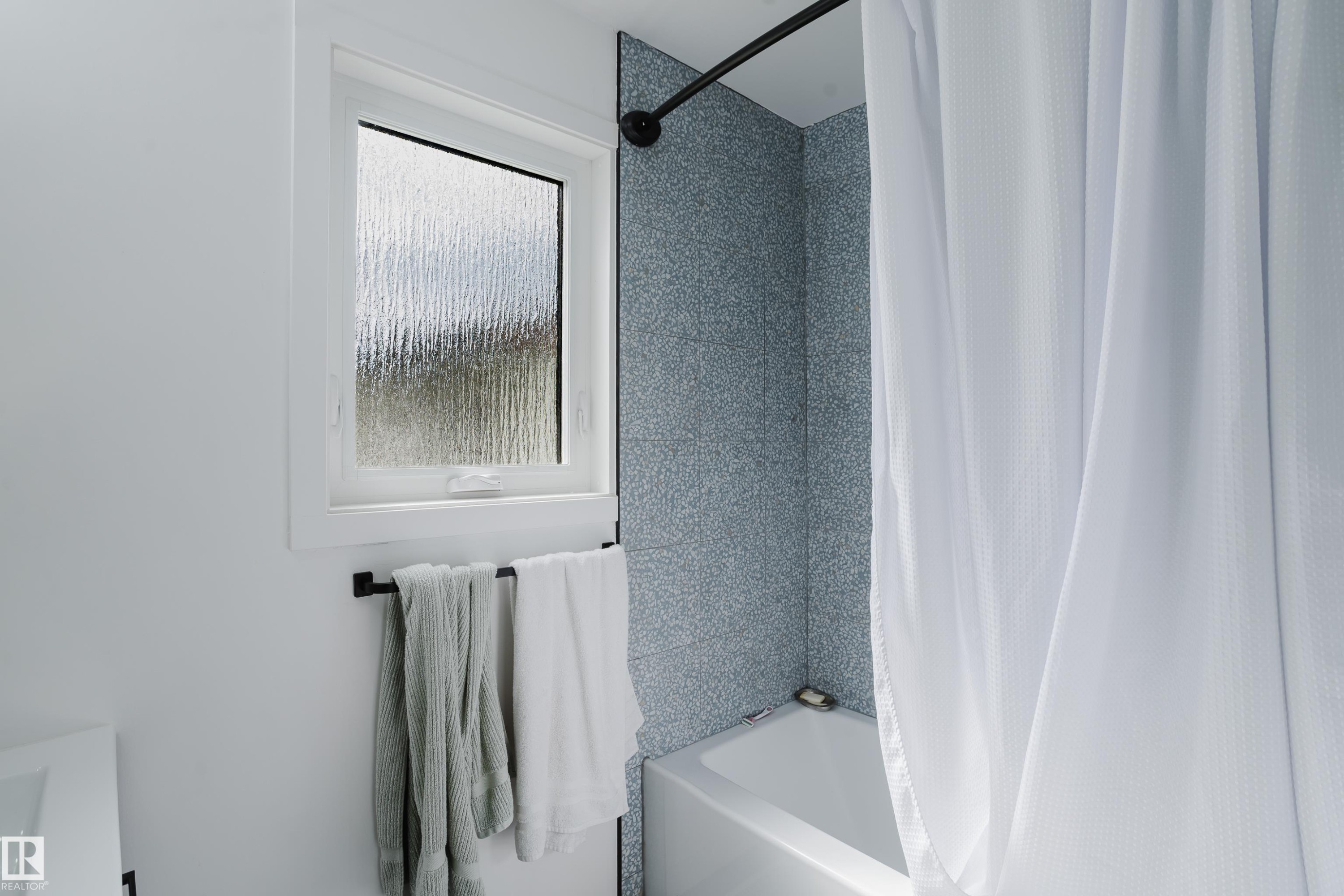 White bathtub with a curved shower rod, patterned blue-gray wall tile, and a privacy window - 9059 52 Street Nw, Edmonton, AB - Indoor Photo Showing Bathroom