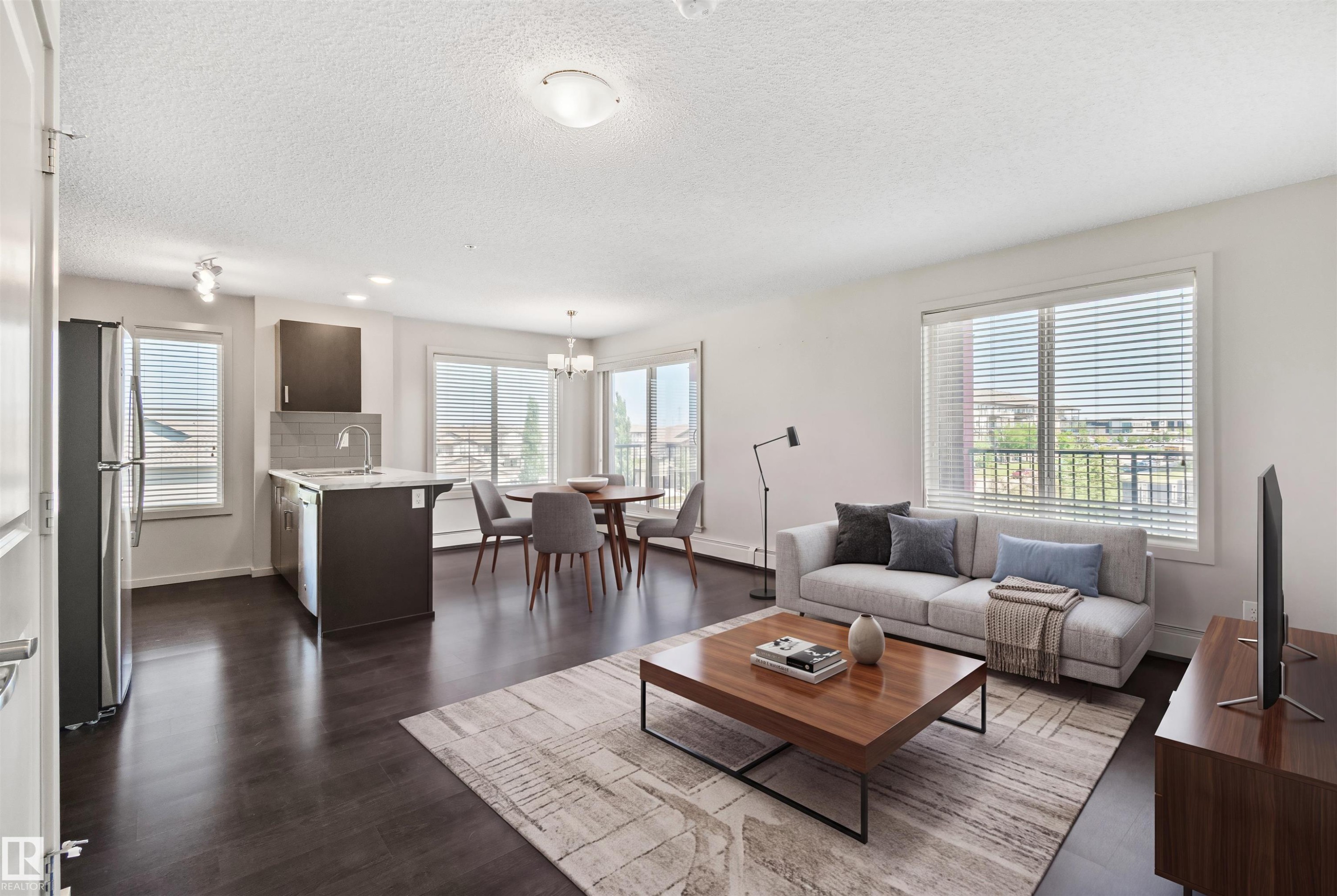 Open-concept living area featuring dark wood-finish flooring, white walls, and large windows with horizontal blinds - #318 5521 7 Av Sw Sw, Edmonton, AB - Indoor Photo Showing Living Room