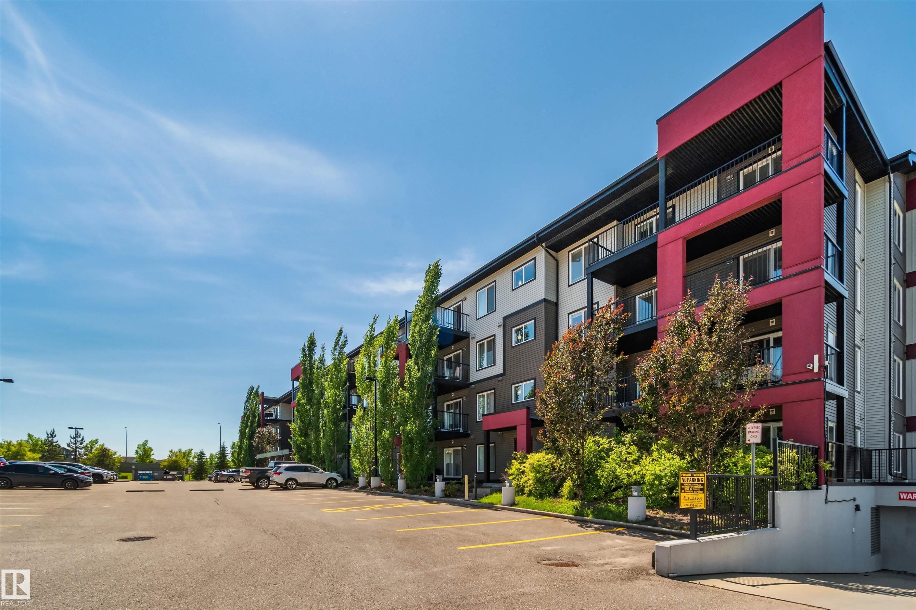 Multi-story building featuring modern red and gray exterior paneling, private balconies, and a paved parking lot with designated spaces - #318 5521 7 Av Sw Sw, Edmonton, AB - Outdoor