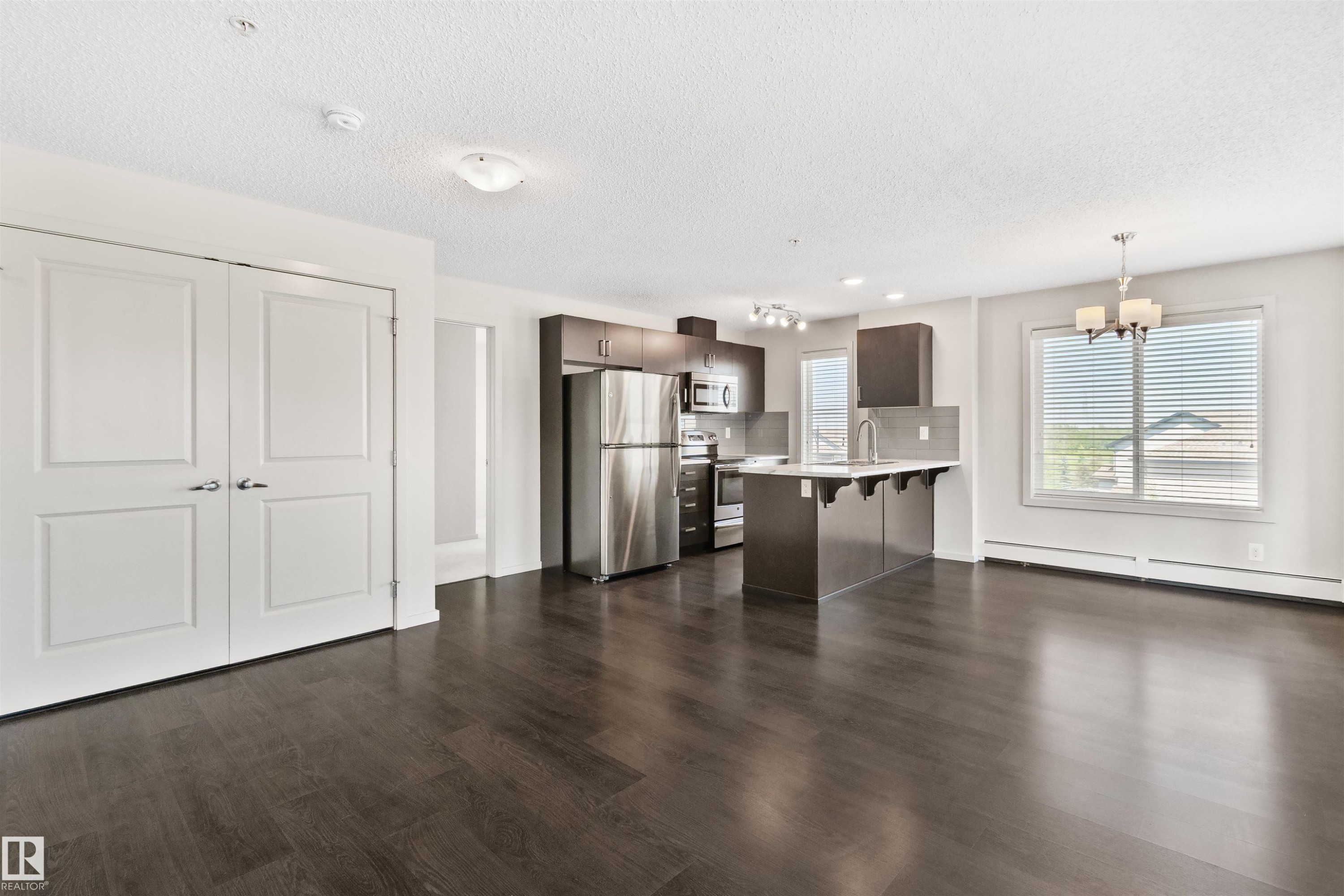 Open-concept living area with dark wood-finish flooring, white walls, and a kitchen featuring stainless steel appliances, dark cabinetry, and an island with bar seating - #318 5521 7 Av Sw Sw, Edmonton, AB - Indoor Photo Showing Kitchen