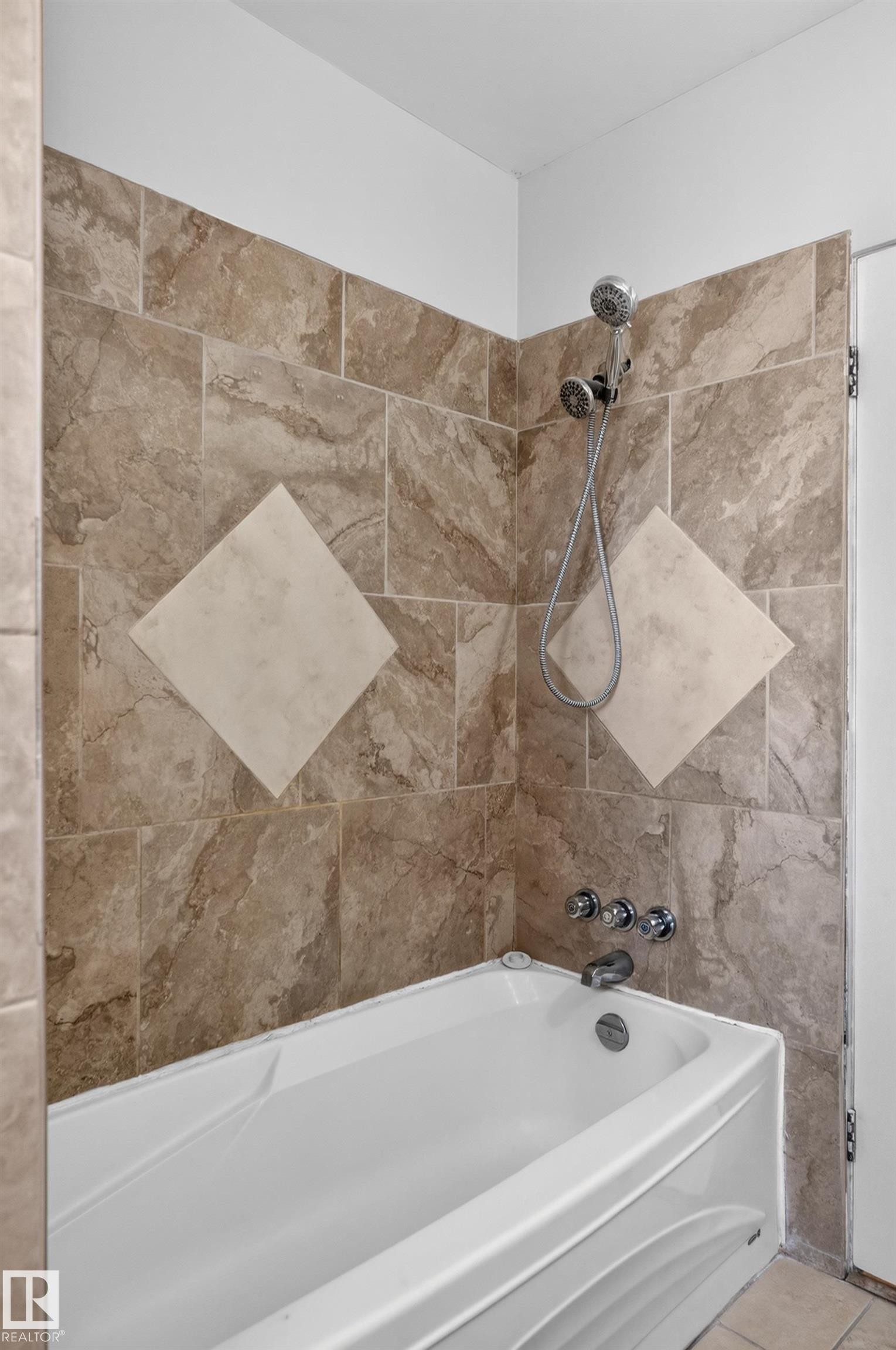 White bathtub with polished chrome fixtures, surrounded by neutral-toned tiled walls featuring a decorative diamond pattern - 13214 123 Street Nw, Edmonton, AB