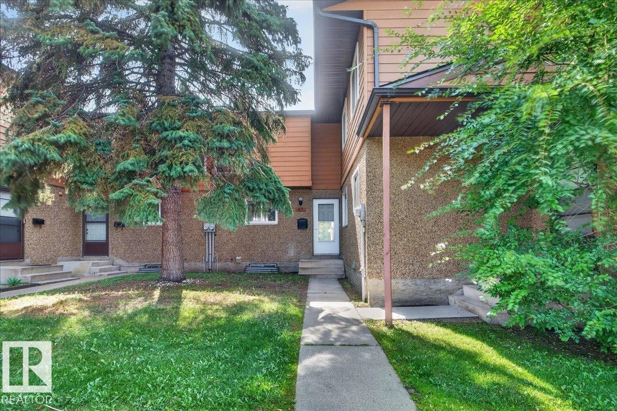 Exterior featuring a concrete walkway, established front lawn, mature evergreen tree, and stucco facade with a covered entry - #47 3812 20 Av Nw, Edmonton, AB