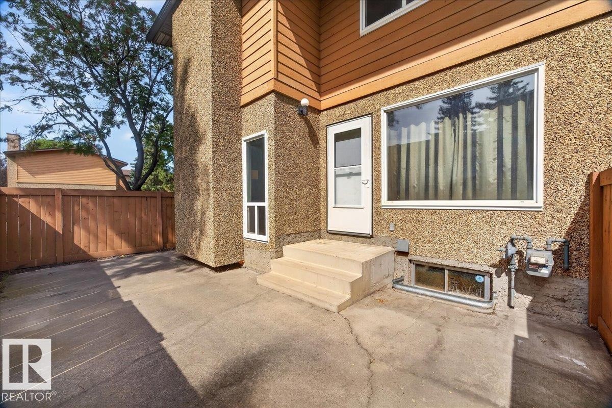 Concrete patio area featuring a wooden privacy fence, textured exterior siding, and multiple windows - #47 3812 20 Av Nw, Edmonton, AB
