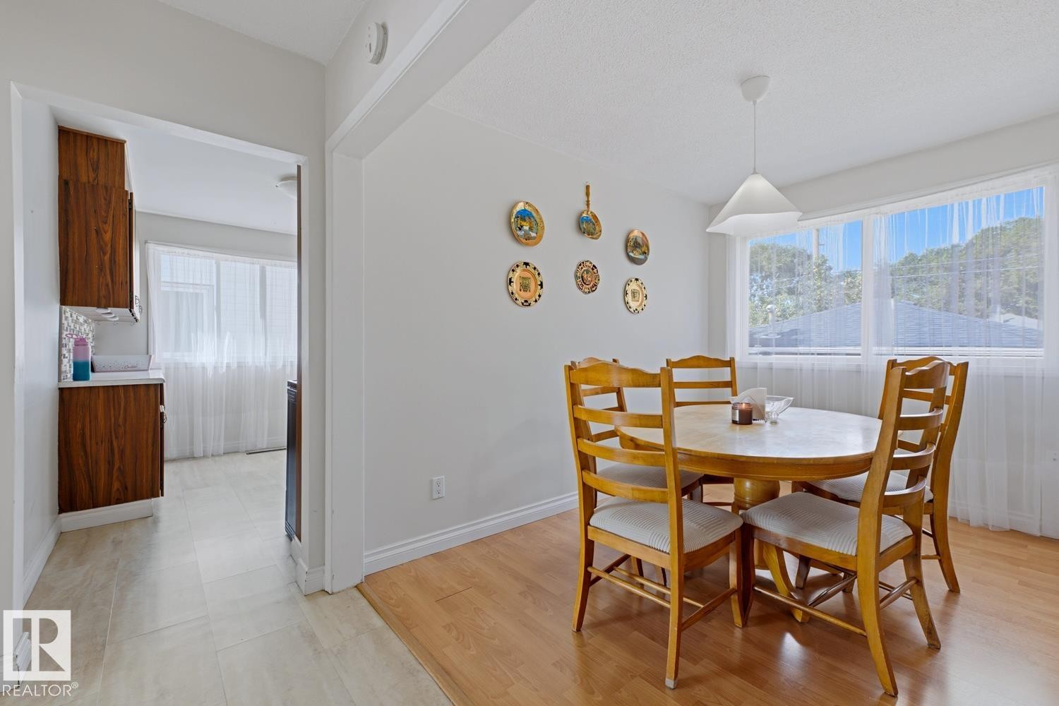 Bright dining area featuring warm wood-finish flooring, a round wood dining table with ladder-back chairs, and expansive windows with natural light - 9740 63 Avenue Nw, Edmonton, AB