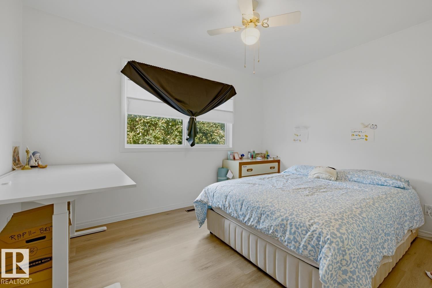Bright secondary bedroom featuring wood-finish flooring, a large operable window with motorized blackout shade, white painted walls, and built-in desk workspace with integrated storage - 9740 63 Avenue Nw, Edmonton, AB