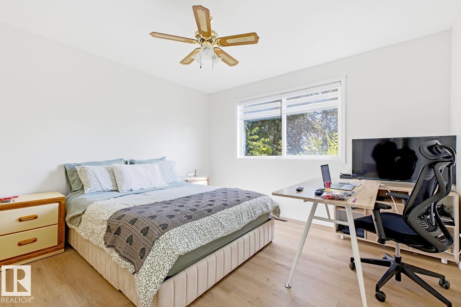 Bright bedroom with wood-finish flooring, large dual-pane windows with natural light, ceiling fan with brass finish, wood nightstand with drawer storage, and dedicated workspace with desk and ergonomic seating - 9740 63 Avenue Nw, Edmonton, AB