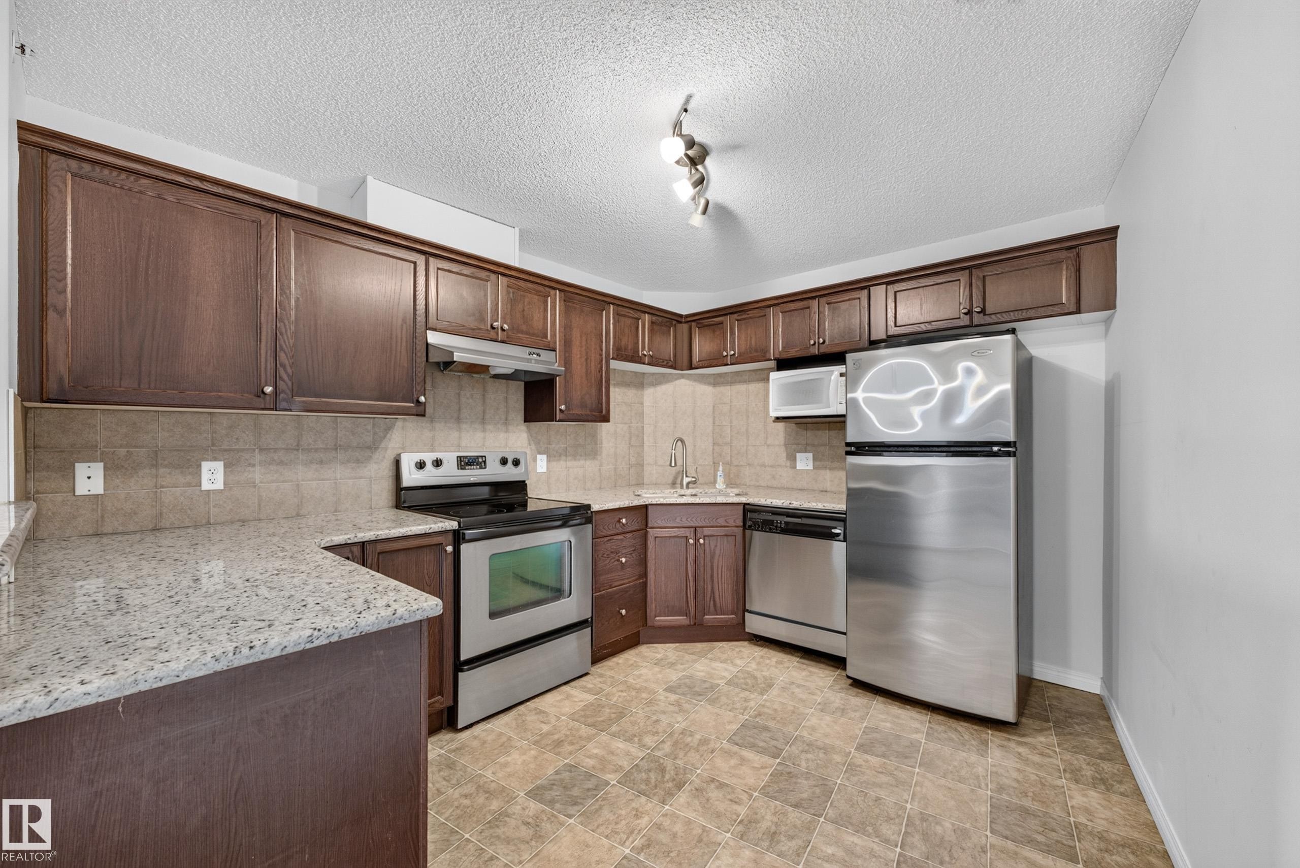 Kitchen featuring wood-finish cabinetry, light-colored countertops, stainless steel appliances, a tile backsplash, and patterned flooring - #6430 7331 South Terwillegar Dr Nw, Edmonton, AB