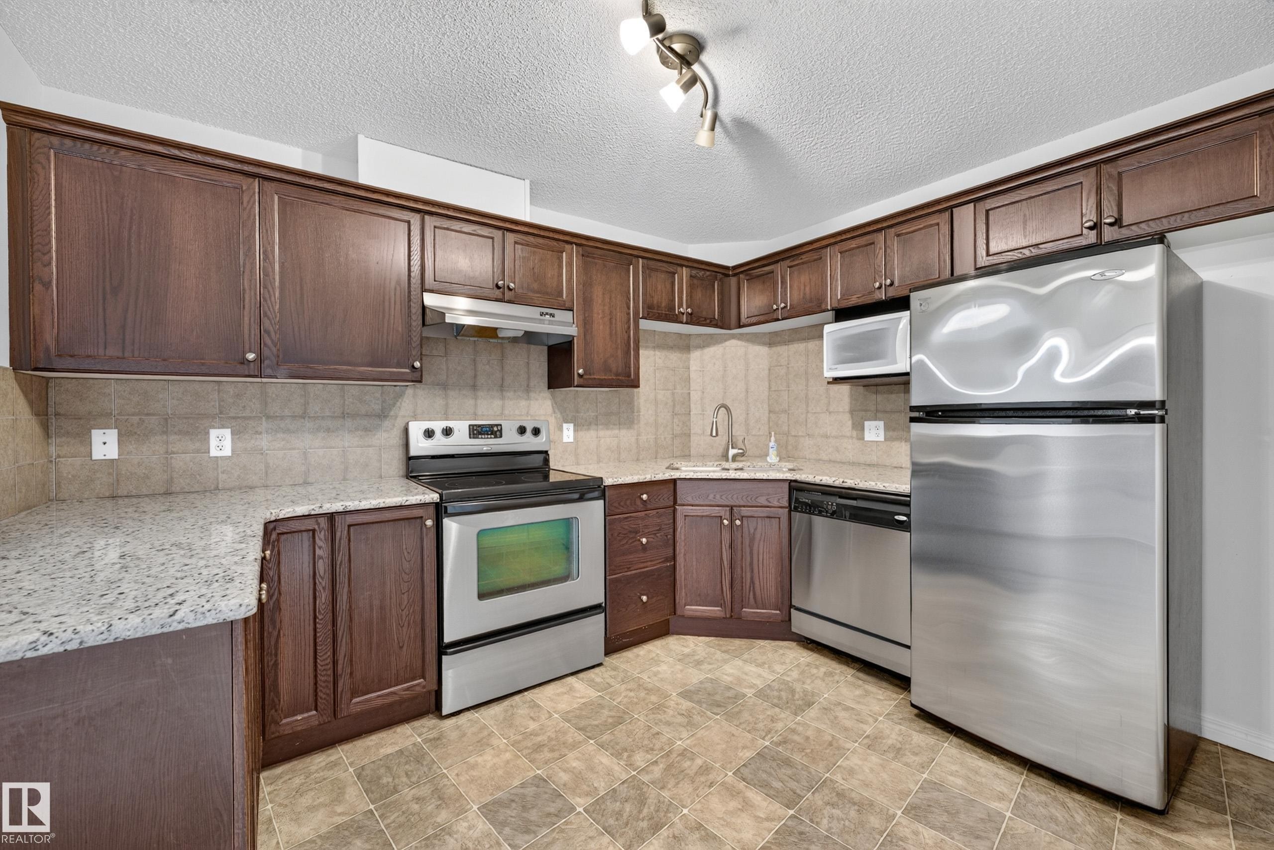 Kitchen featuring dark wood-finish cabinetry, light-toned stone-finish countertops, stainless steel appliances, and a tiled backsplash - #6430 7331 South Terwillegar Dr Nw, Edmonton, AB