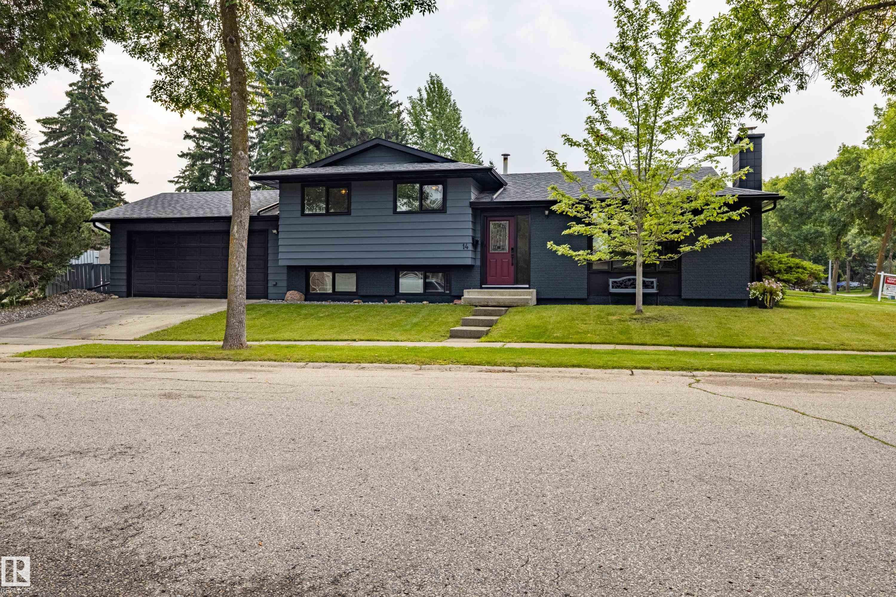Modern Contemporary home featuring a striking black exterior with clean architectural lines, attached two-car garage, and a professionally landscaped front yard with mature trees and manicured lawn - 14 Brunswick Crescent, St. Albert, AB