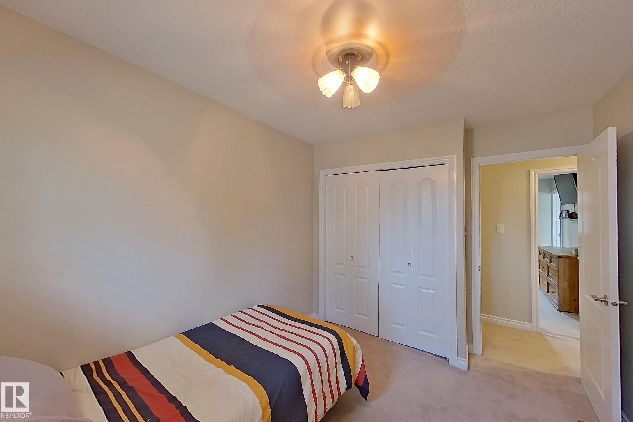 Bright secondary bedroom featuring neutral carpeting, a ceiling-mounted light fixture with three-bulb configuration, and dual closets with white panel doors - 14 Brunswick Crescent, St. Albert, AB