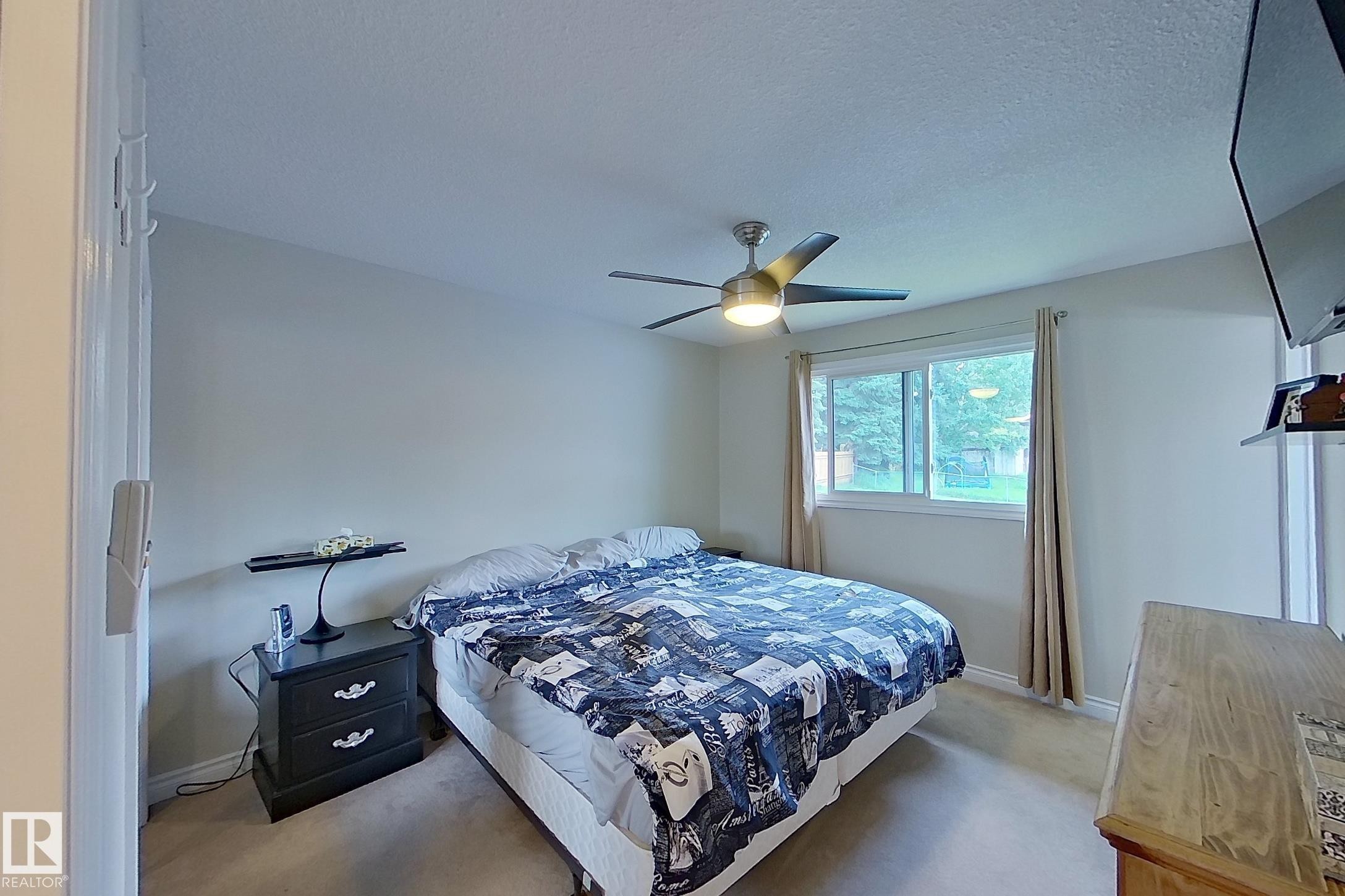 Bright secondary bedroom featuring neutral carpeting, large window with natural light, ceiling fan with integrated lighting, and black nightstand with storage drawers - 14 Brunswick Crescent, St. Albert, AB